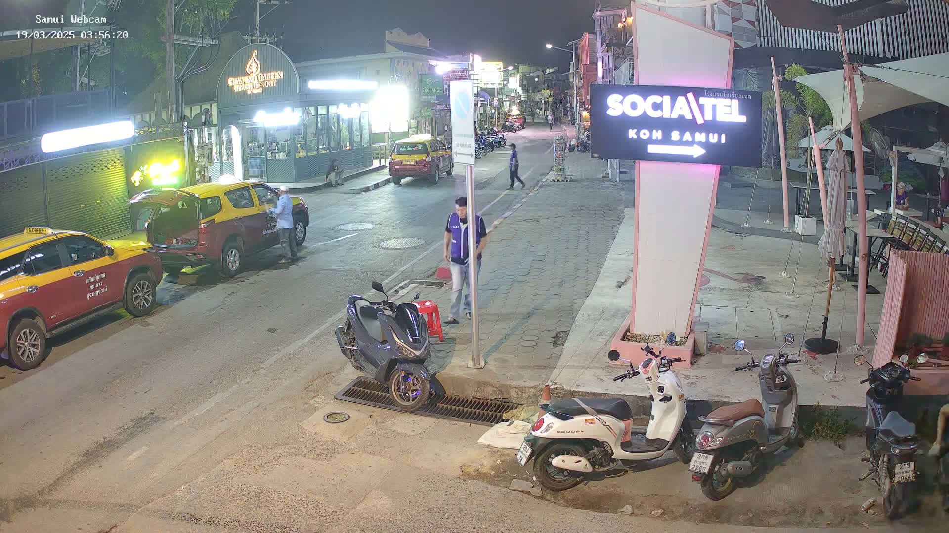 A nighttime street scene shows several people and parked vehicles, including taxis and motor scooters, near a restaurant.