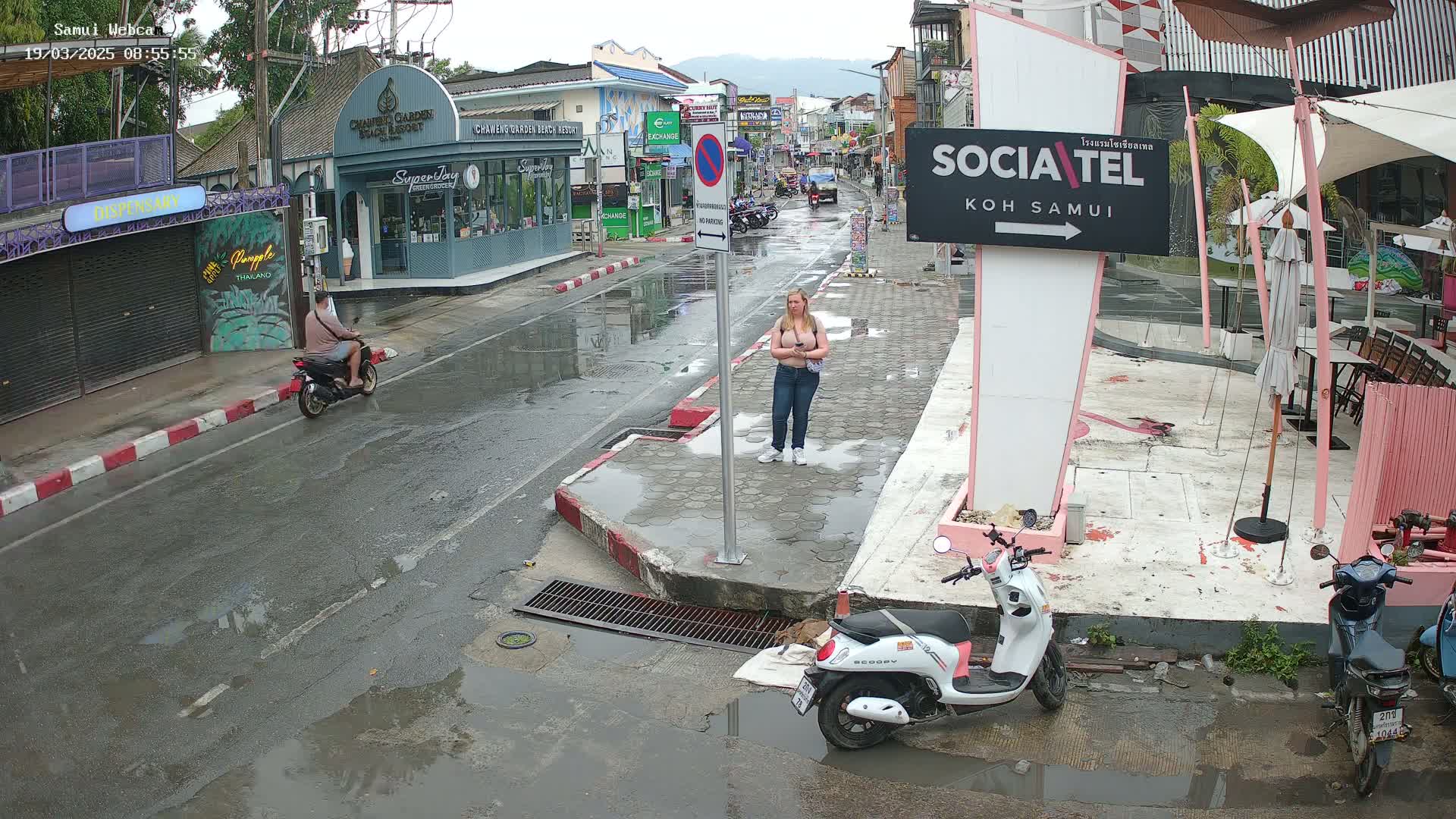A wet street scene shows a woman standing on a sidewalk next to parked motorbikes, with shops and buildings lining the street under an overcast sky.