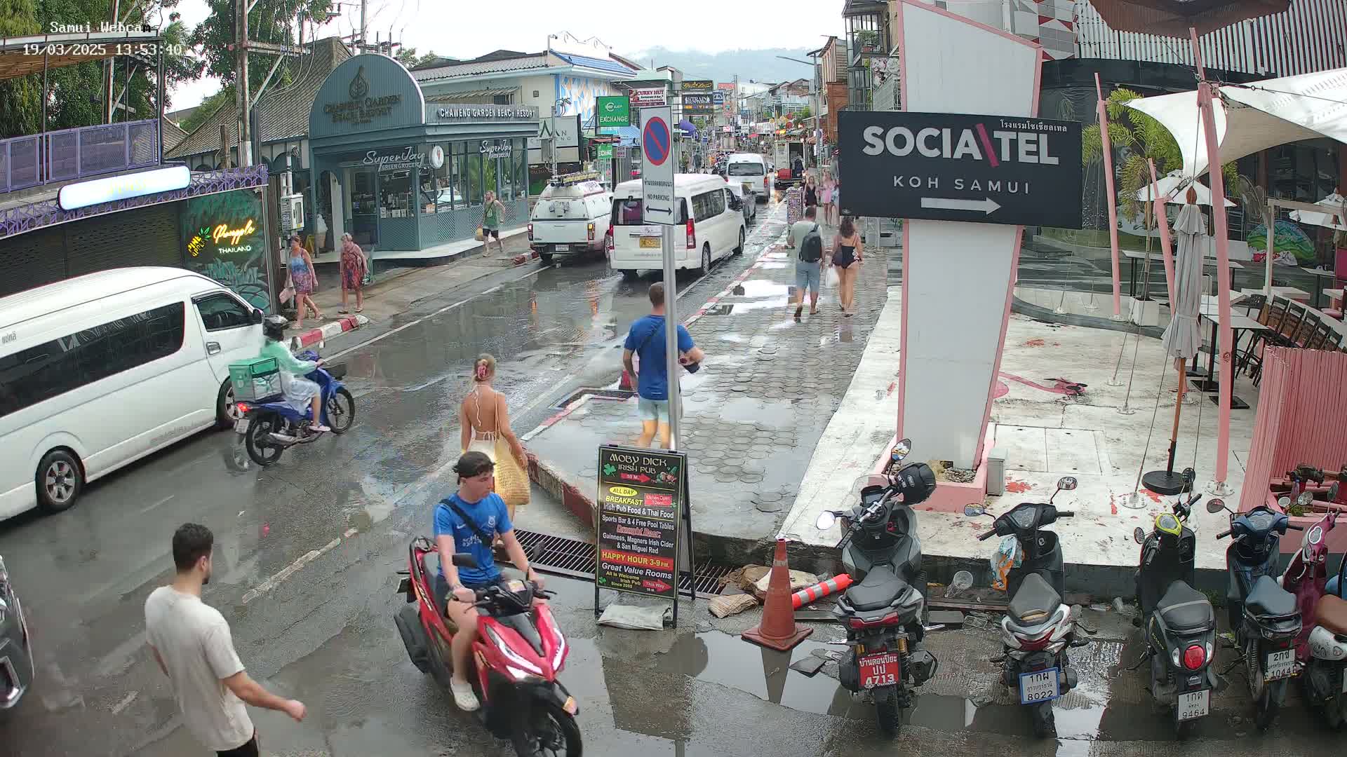 A wet, paved street scene shows people walking and riding scooters amidst parked vehicles and outdoor seating areas under an overcast sky.
