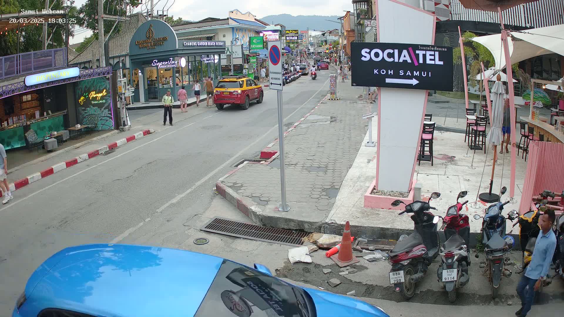 A street scene in Koh Samui shows several pedestrians, cars, and parked motorbikes under partly cloudy skies, with businesses lining the street.