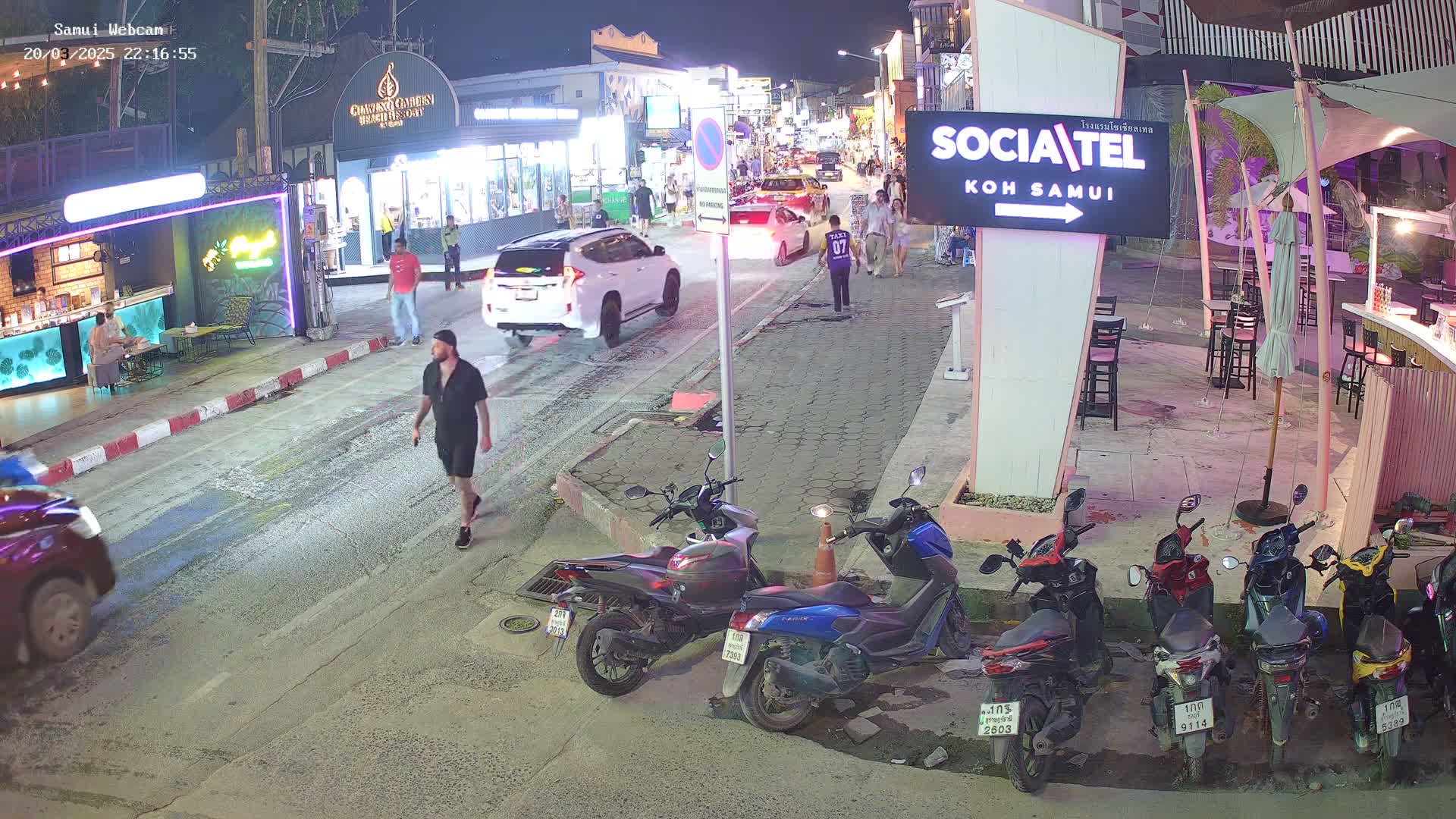 A nighttime street scene shows a man walking on a paved road lined with shops and parked motorbikes, with cars driving by under ambient lighting.