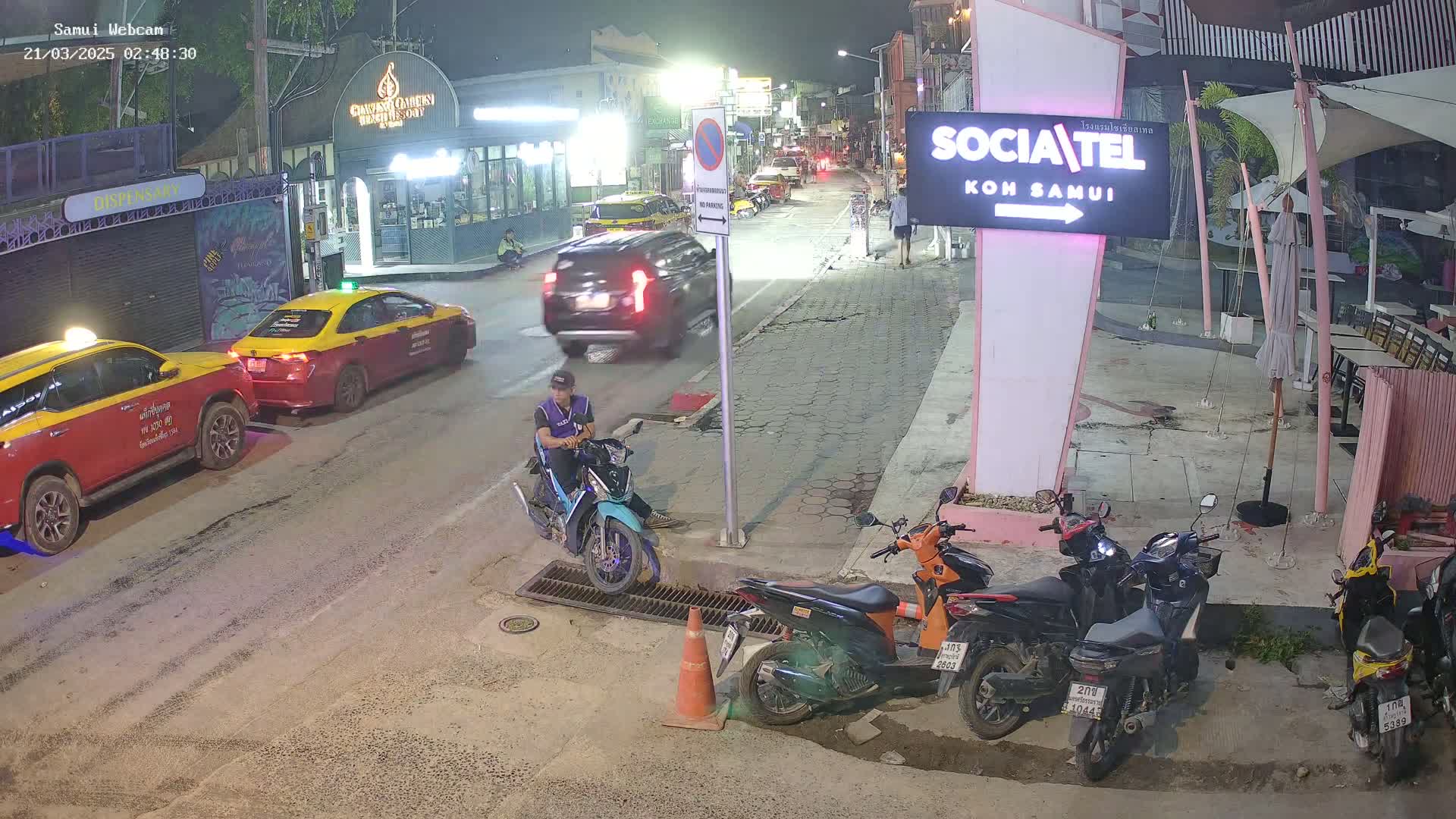 At night, a man sits on a teal scooter parked on the side of a street, with several parked motorcycles nearby, two taxis, and other vehicles driving past.