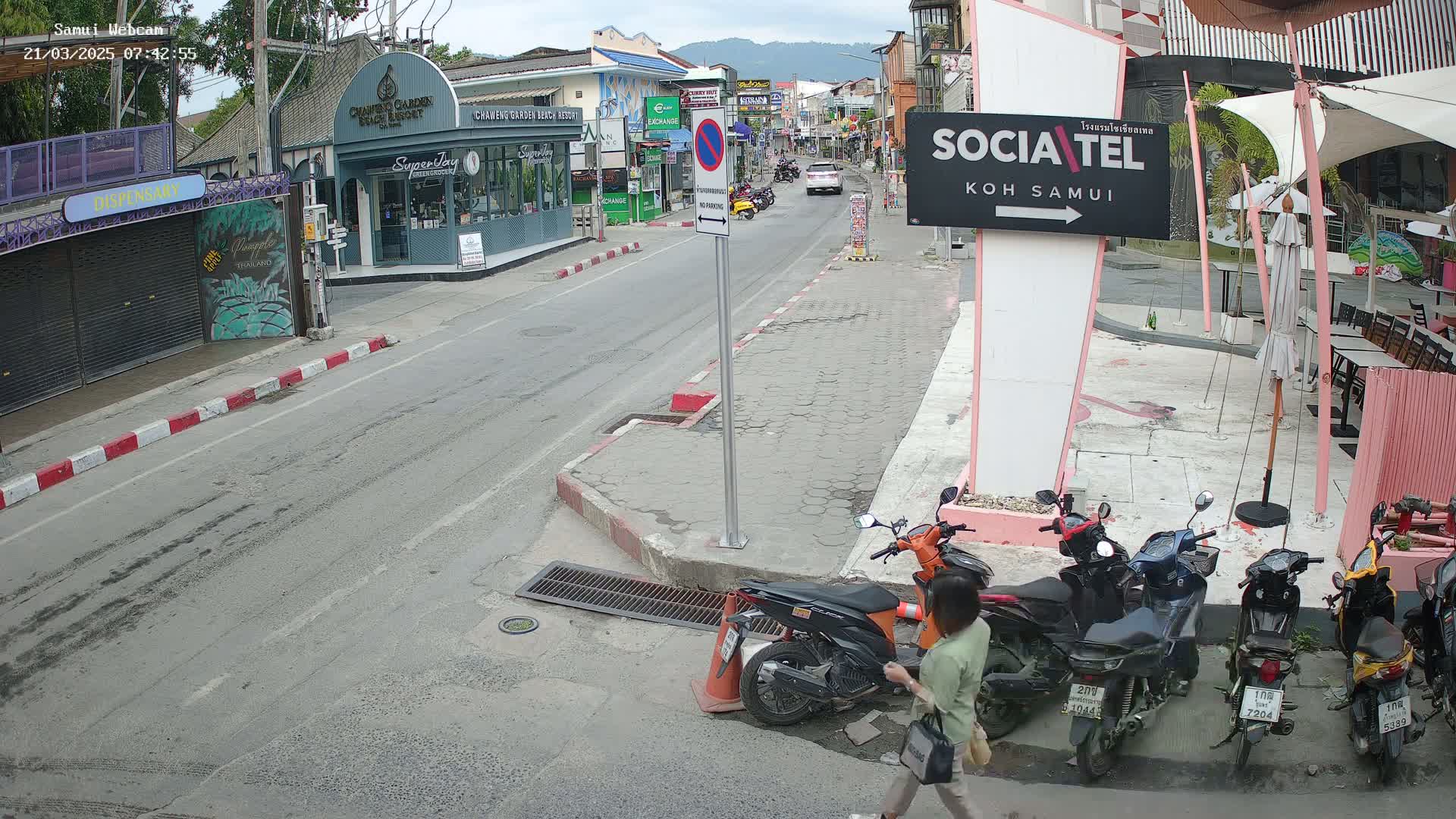 A street scene on a partly cloudy day shows parked motorbikes and cars near shops and restaurants.