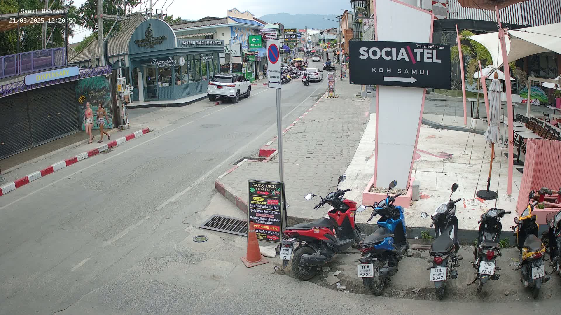 A sunny street scene shows two women walking on a sidewalk, several parked motorbikes, and cars driving on a road lined with shops and buildings.