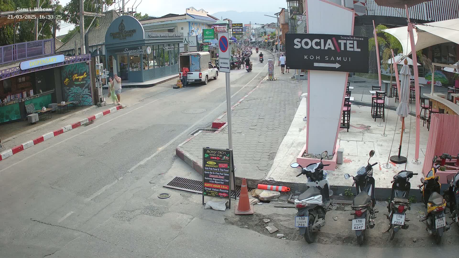 A street scene in bright daylight shows a road with pedestrians, parked motorbikes, and several businesses, including a restaurant with outdoor seating.
