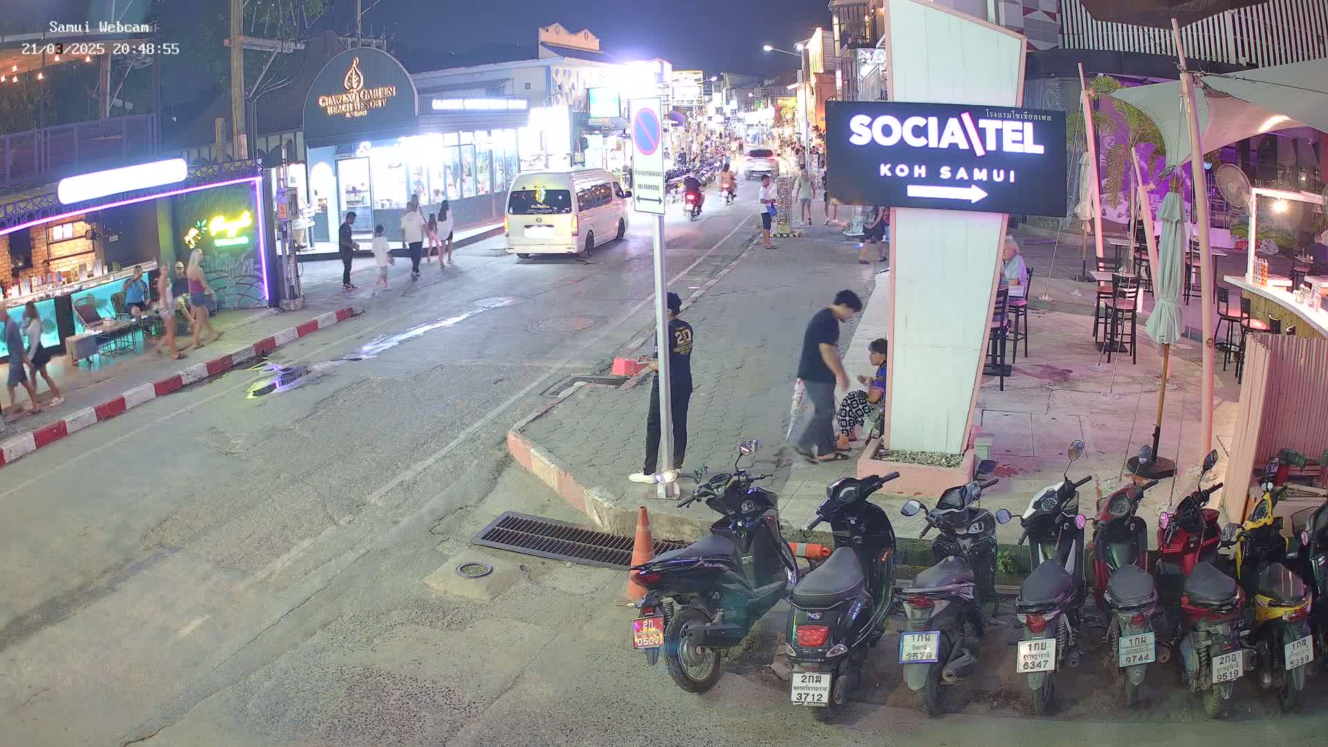 At night, a street scene shows pedestrians walking past shops and restaurants, with several parked motorbikes and a van in the background.