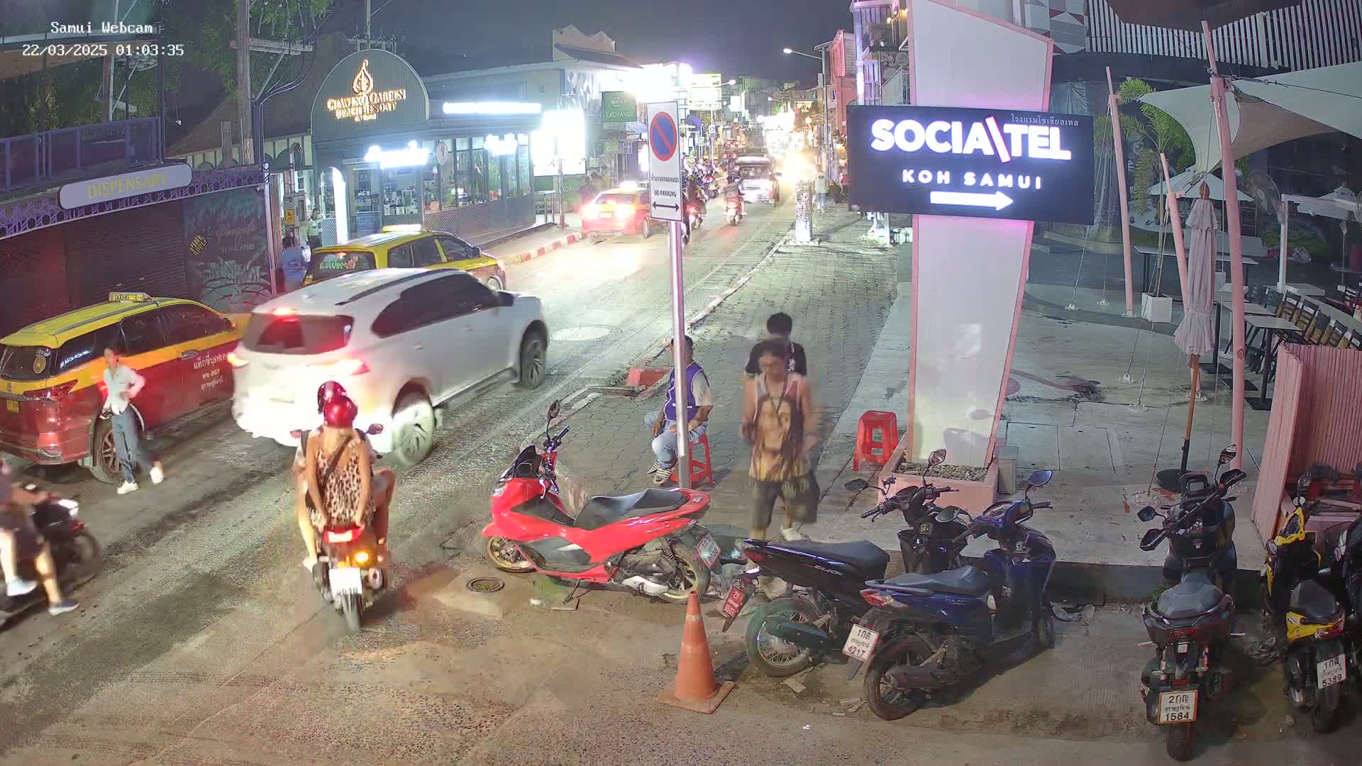 A nighttime street scene shows several cars and motorcycles parked and moving on a roadway, with people walking along the sidewalks near shops and restaurants.