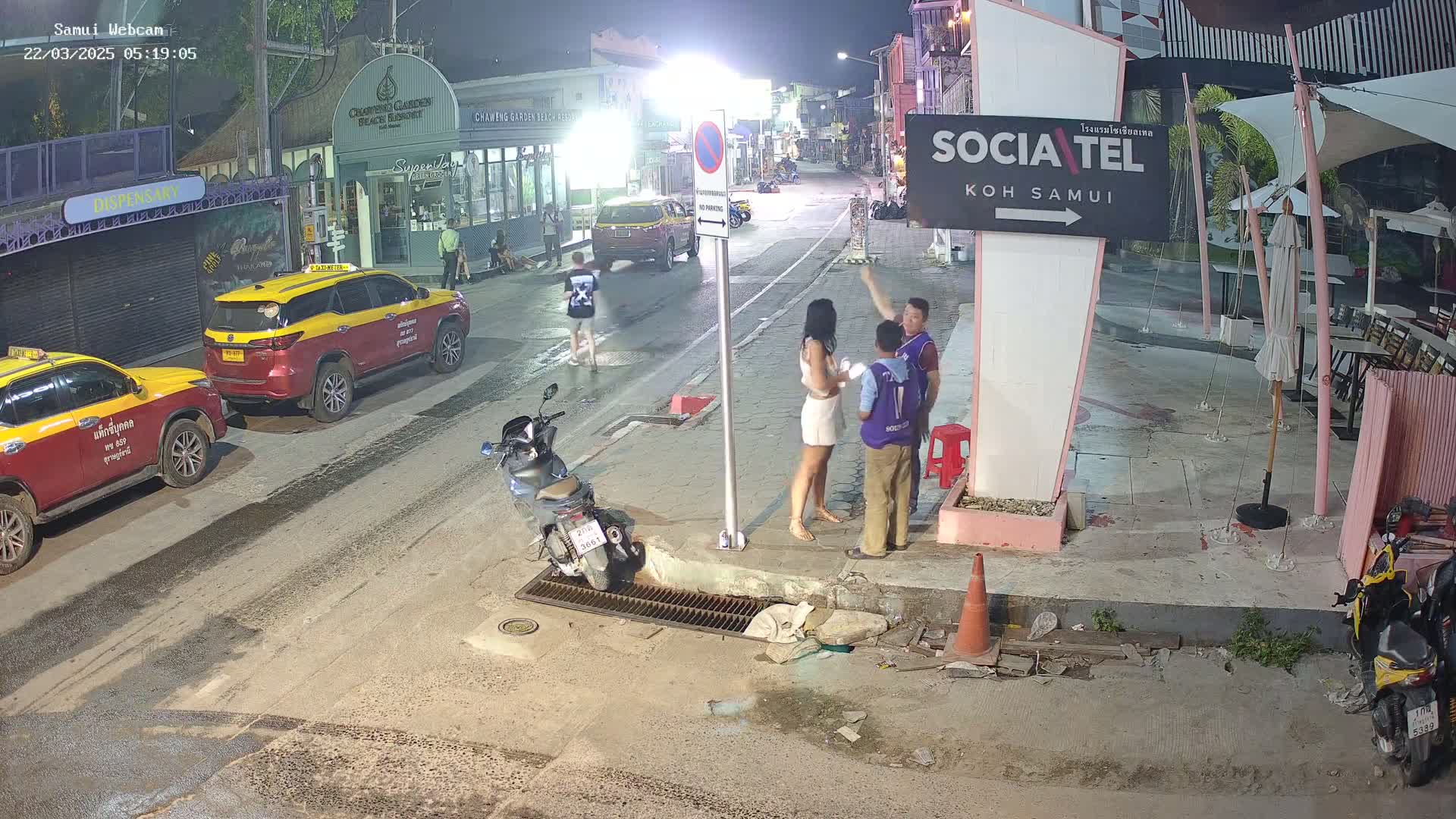 At night, three people converse near a pink sign on a street with parked taxis and motorbikes under the light of street lamps.
