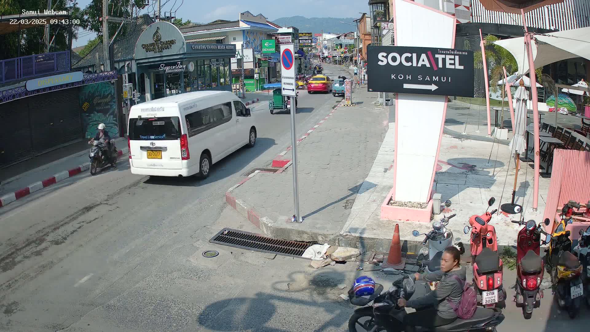 A sunny street scene shows several motorbikes parked, a person on a motorbike, a white van, and other vehicles driving down a road past shops and restaurants.