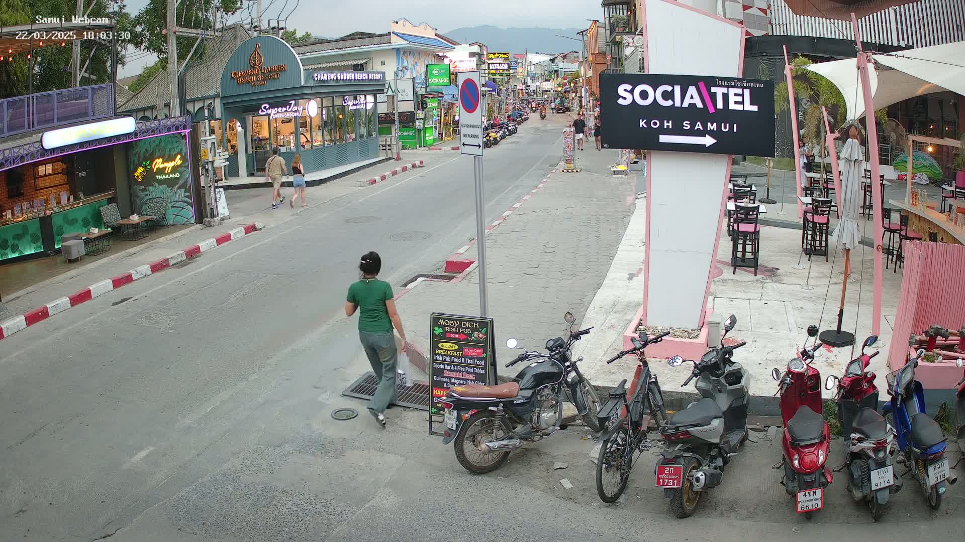 A street scene shows a woman walking on a road with parked motorbikes and shops lining either side, under an overcast sky.