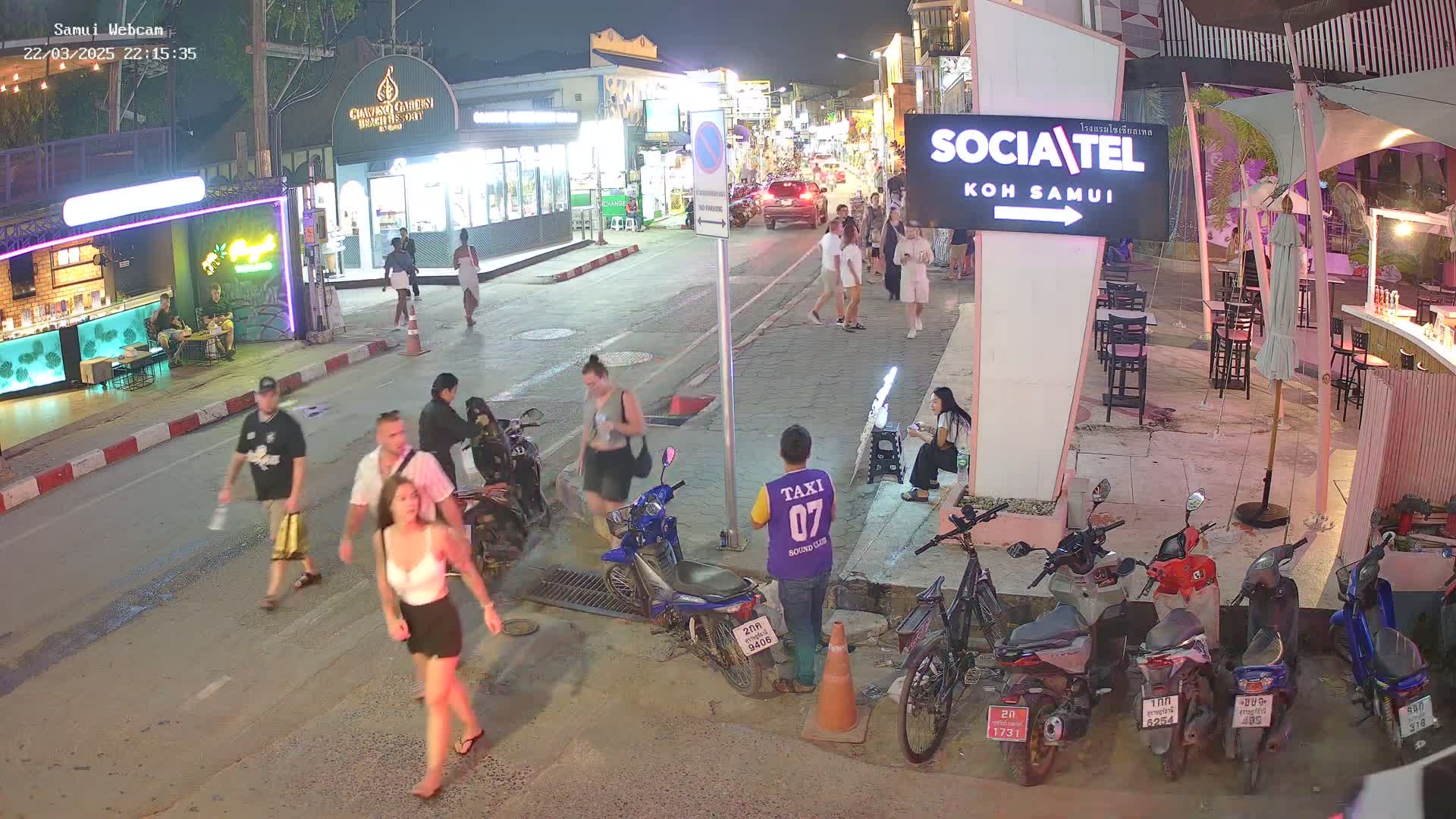 A nighttime street scene shows pedestrians and parked motorcycles along a road lined with shops and restaurants under clear skies.