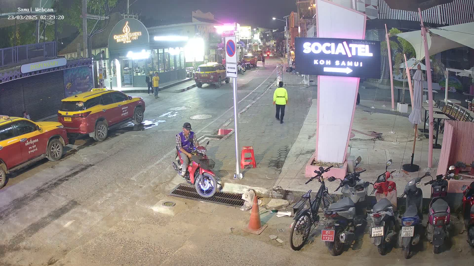 A nighttime street scene shows a person on a red moped, parked taxis, several motorcycles parked at a curb, and a pedestrian walking down a street under dim lighting.