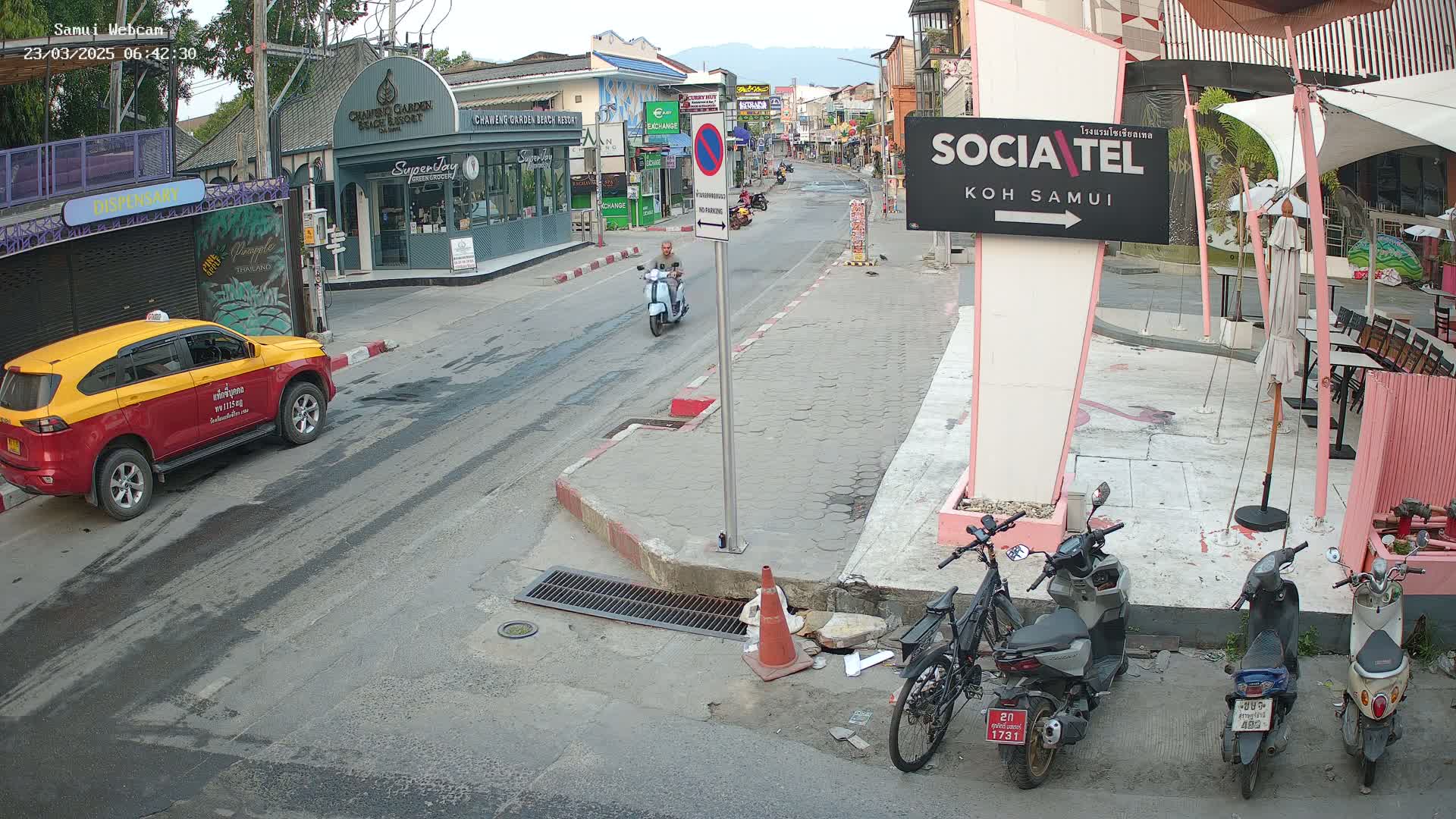 A mostly empty street scene in daytime shows a red and yellow taxi, several motorbikes, and a bicycle parked near a restaurant under a mostly clear sky.