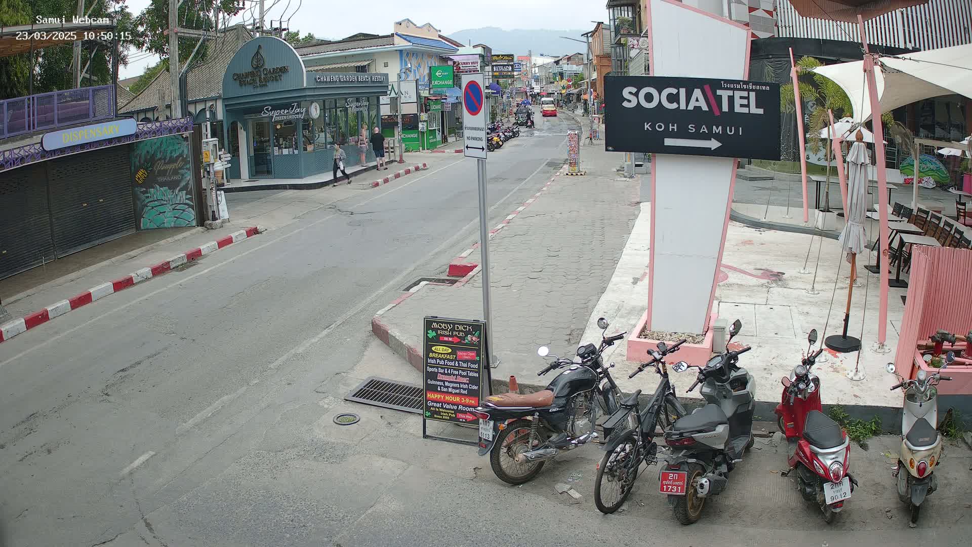 A street scene on a cloudy day shows parked motorcycles and scooters along the roadside near shops and restaurants.