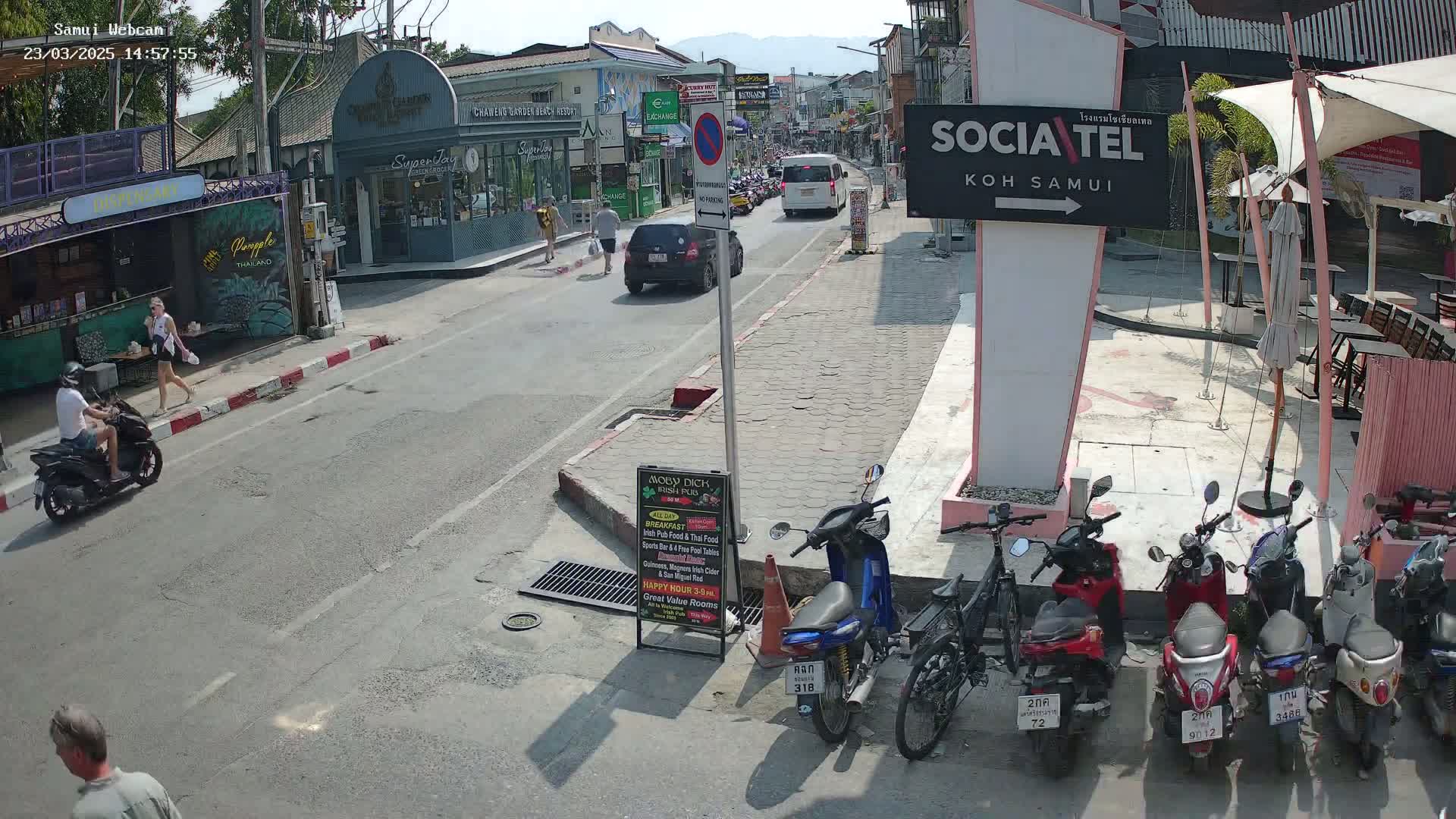 A sunny street scene shows pedestrians and motorbikes near shops and restaurants, with several parked motorbikes in a row.