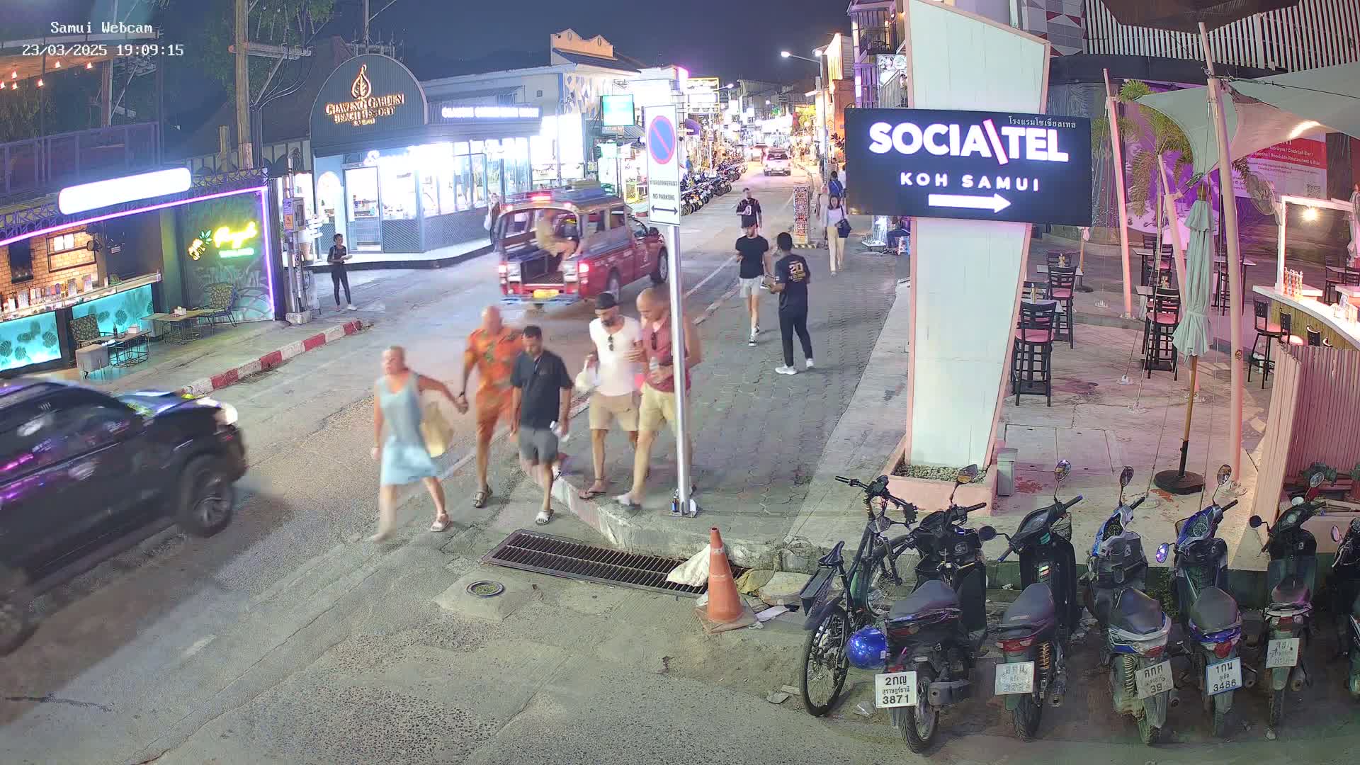 At night, several people walk along a street lined with shops and restaurants, while a red truck and several parked motorbikes are visible.