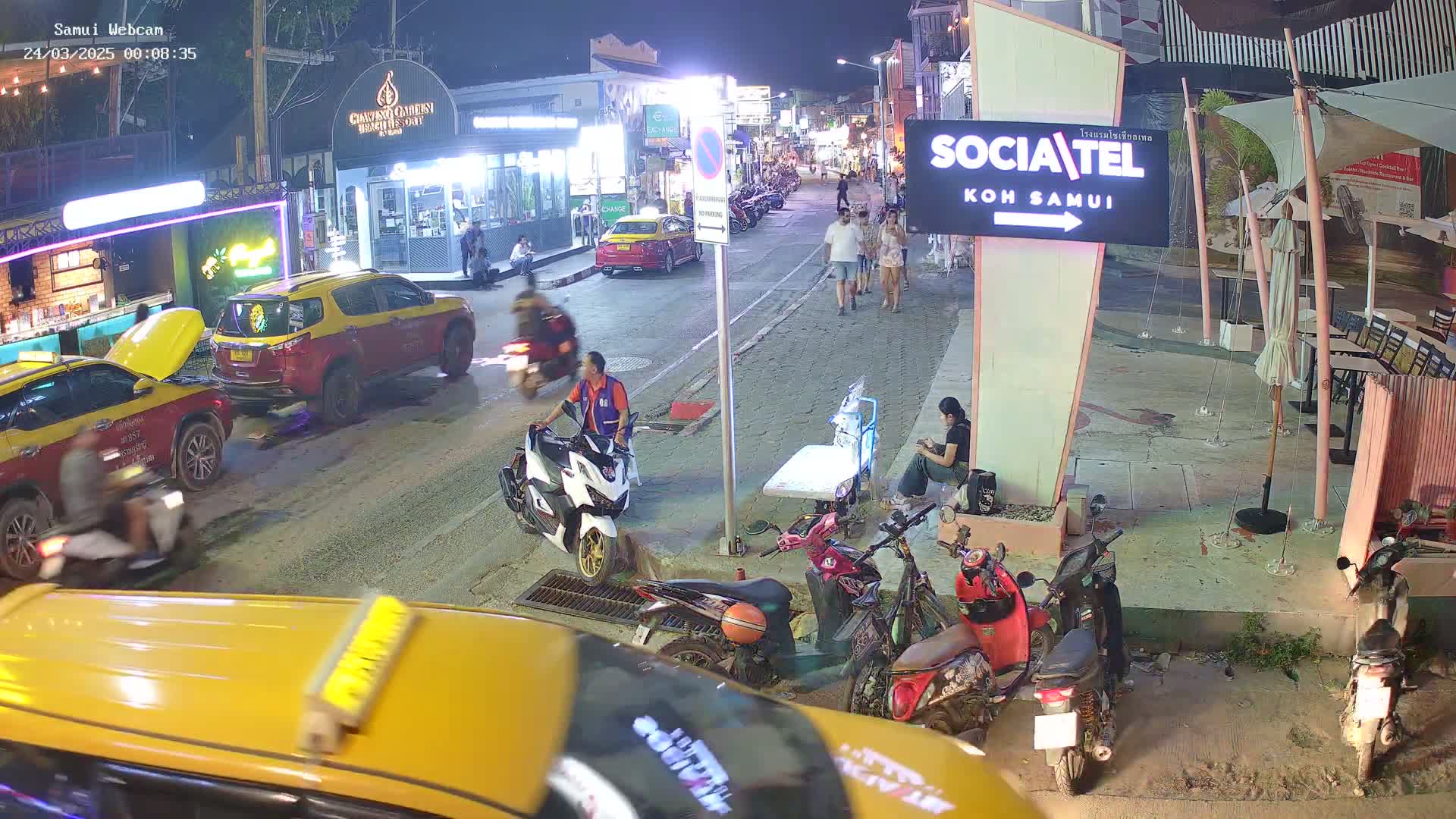 At night, a street scene shows several people walking, motorbikes parked and driven, and taxis stopped in a street with buildings and outdoor seating areas.