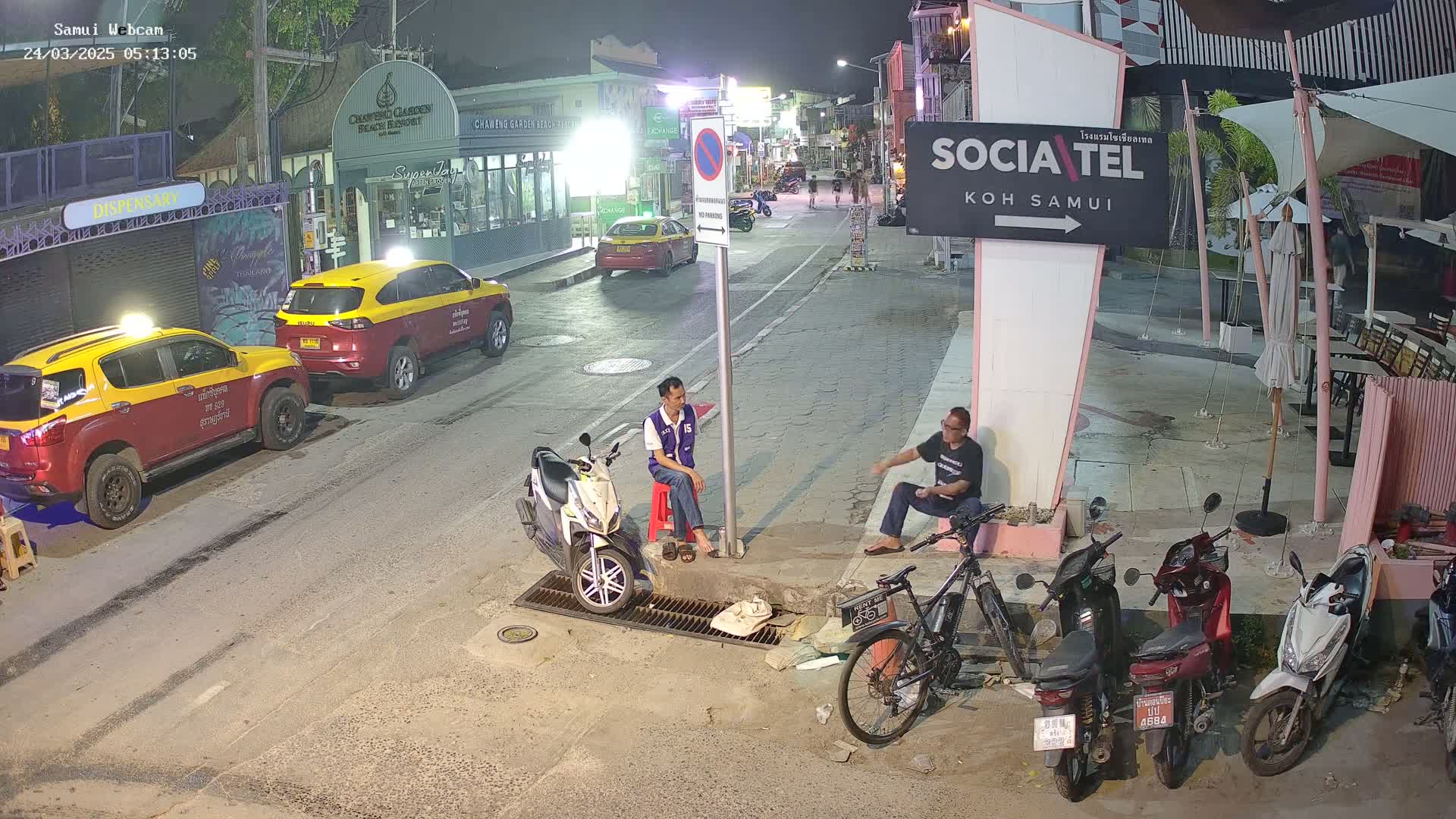 At night, two men sit on a curb next to several parked motorbikes and two taxis, near a street under ambient lighting.