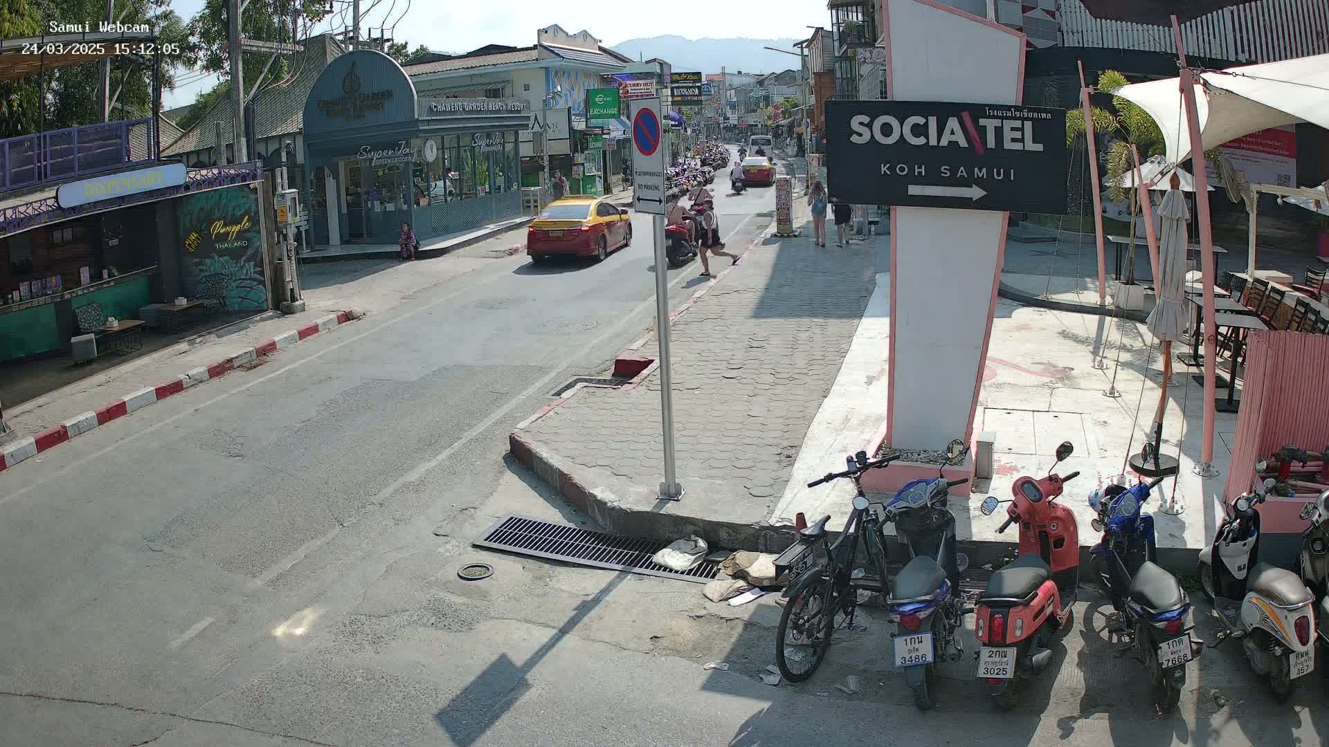 A sunny street scene shows a road with parked motorcycles and cars, pedestrians walking on the sidewalk, and shops along the street.