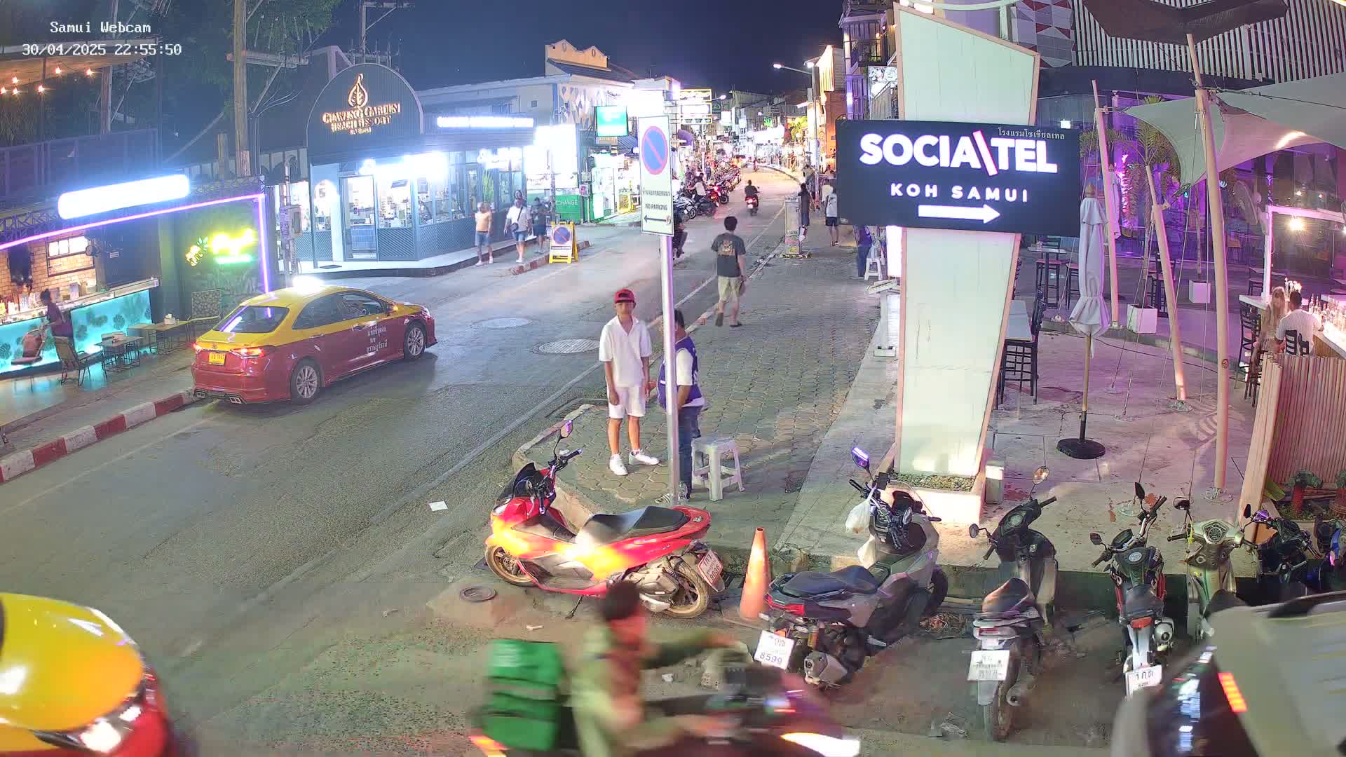 A nighttime street scene shows several parked motorbikes and a taxi, with people walking along a sidewalk lined with shops and restaurants.
