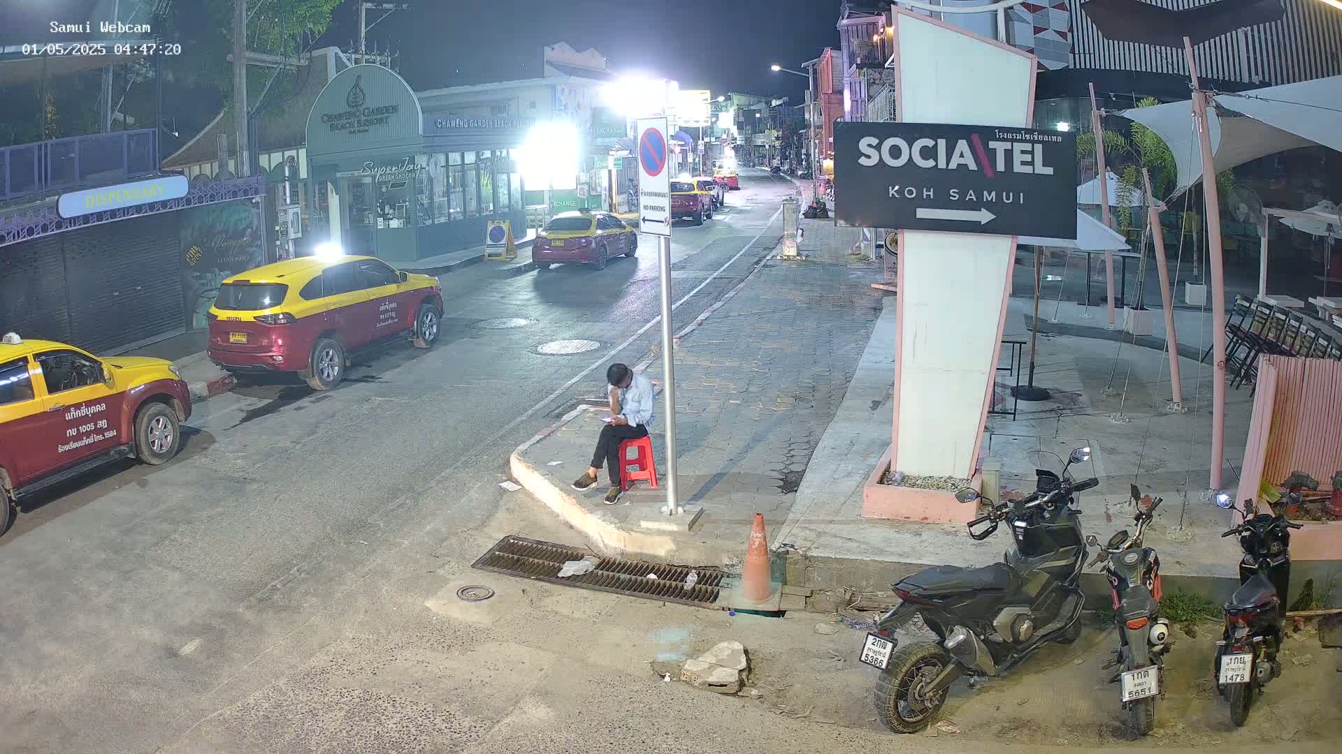 At night, a person sits on a red stool on a street corner next to parked motorcycles, with several taxis and buildings visible in the background under dim lighting.