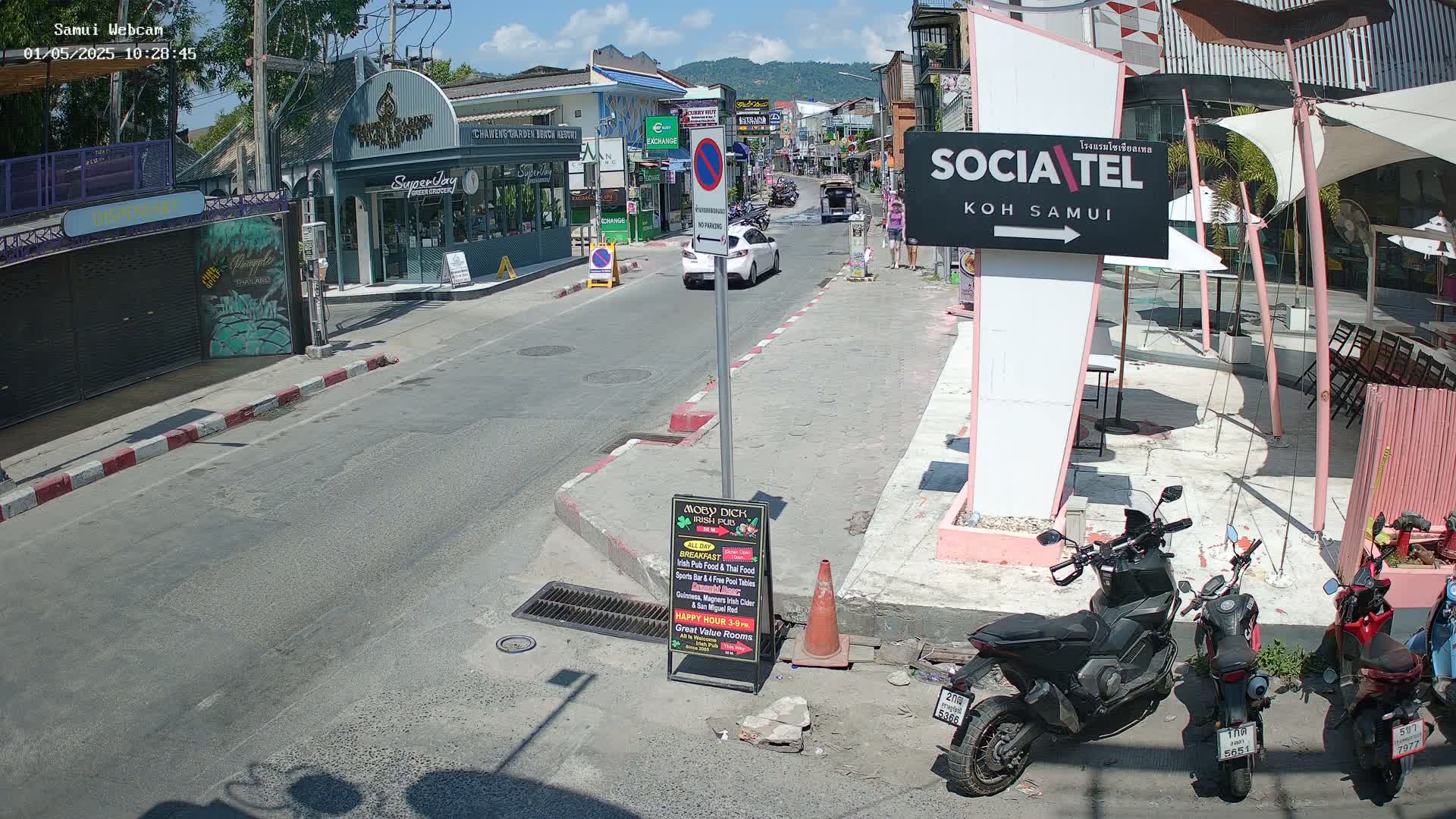 A sunny street scene shows parked motorcycles, cars driving on a road, and various shops and restaurants, all under a bright, clear sky.