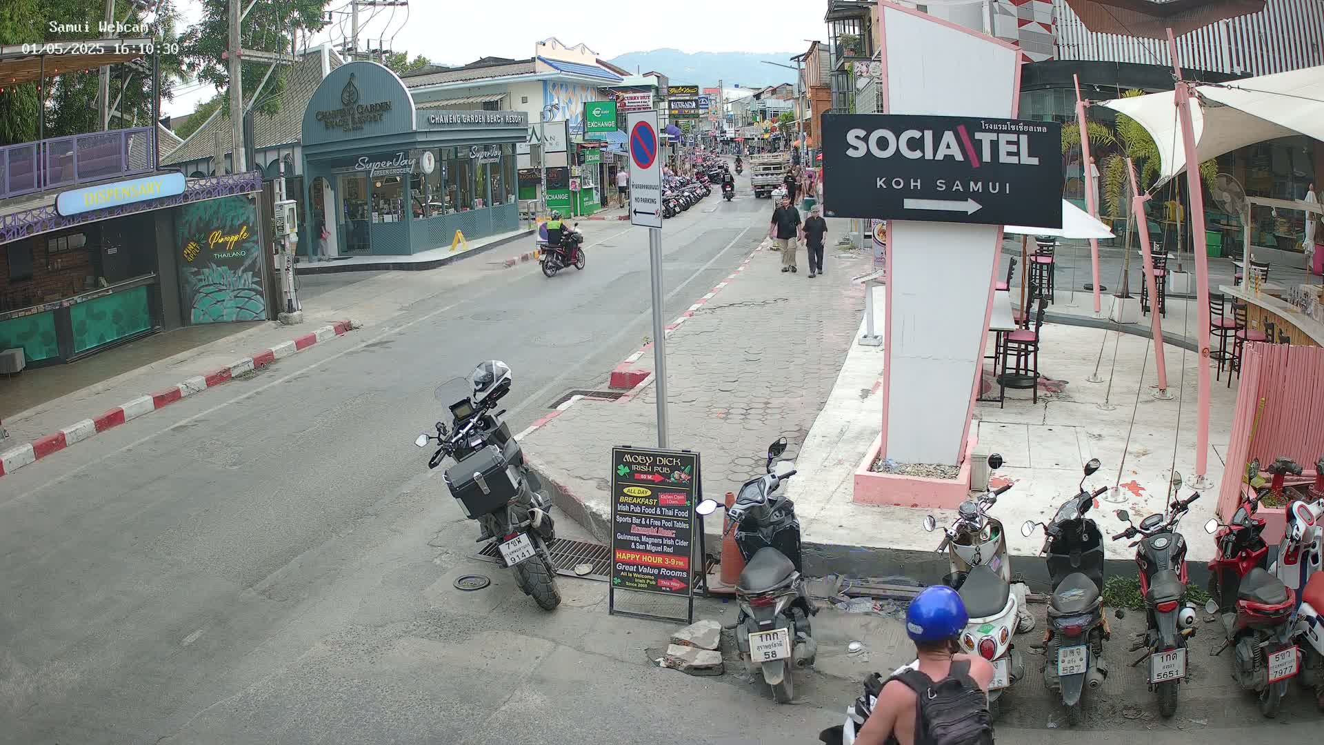 A street scene on a partly cloudy day shows several motorcycles and scooters parked along the side of a road, with people walking and some shops along the street.