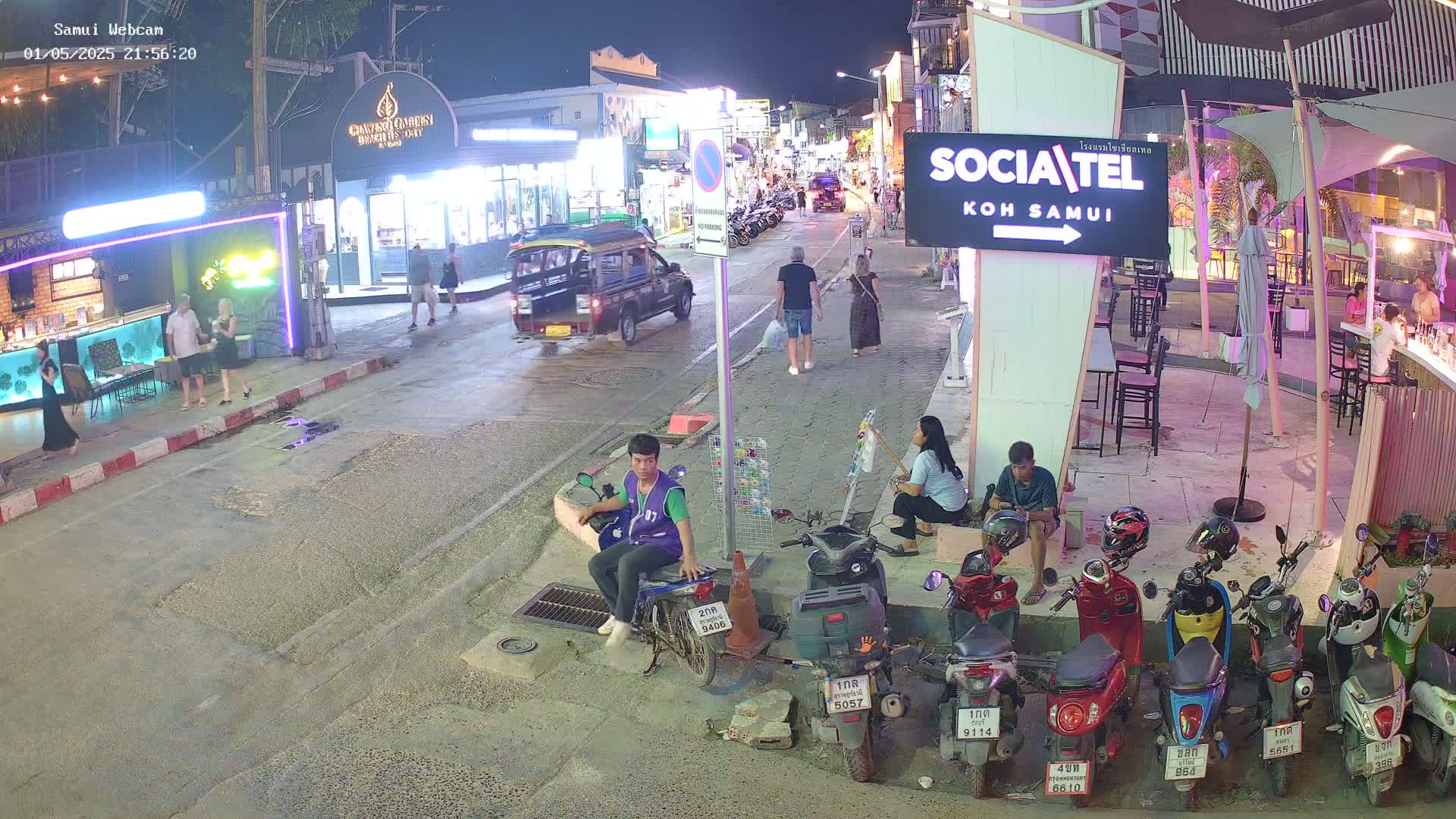 At night, a street scene shows people walking and sitting near parked motorbikes and shops under a partly cloudy sky.