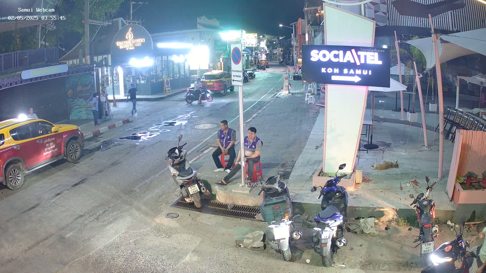 At night, two men sit on stools beside several parked motor scooters on a street with a few cars and pedestrians visible in the background.
