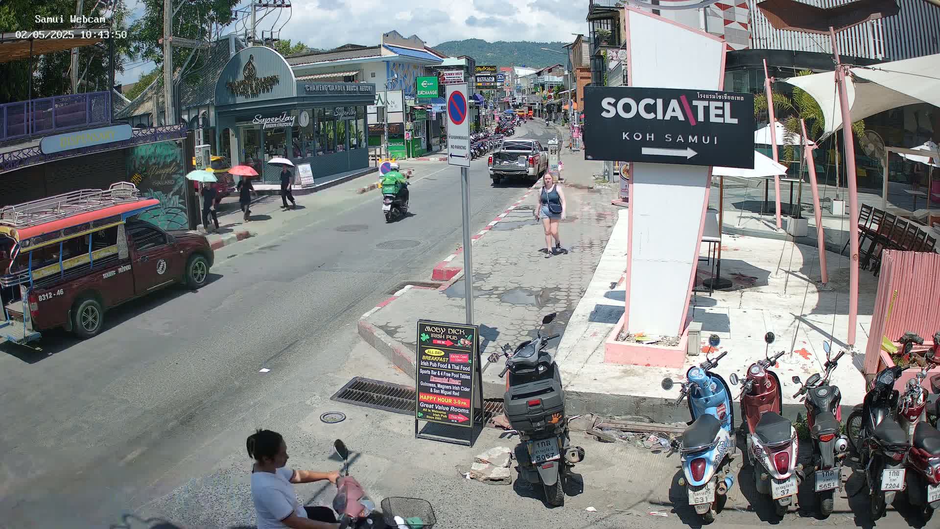 A sunny street scene shows a woman walking, people on motorbikes and in cars, and several parked motorbikes under a pink awning, all in front of various shops and buildings.