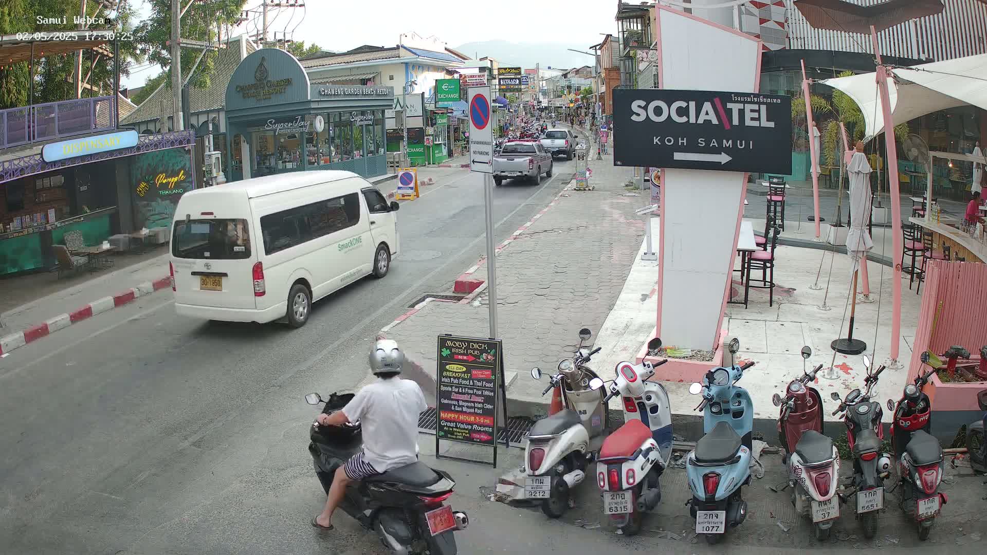 A street scene shows a man on a scooter in front of several parked scooters, a white van, and various shops and restaurants under an overcast sky.