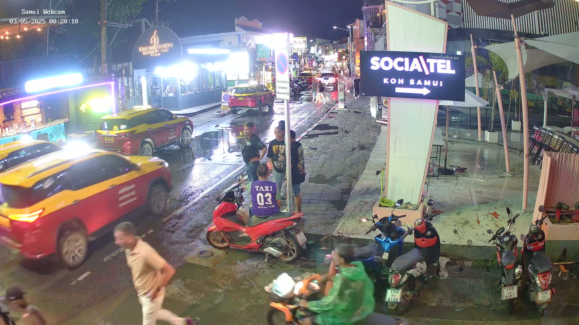 A nighttime street scene shows several people standing near parked motorbikes and taxis, with puddles on a wet roadway under dim lighting.