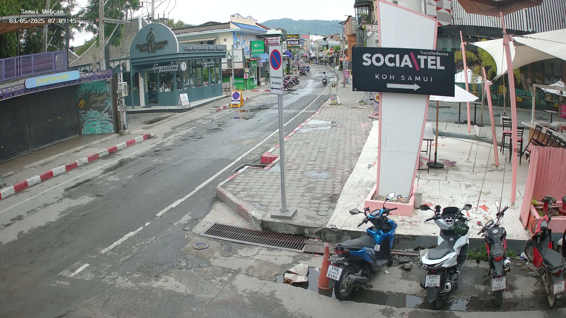 A wet street scene shows several parked motorcycles and shops on a slightly overcast day.