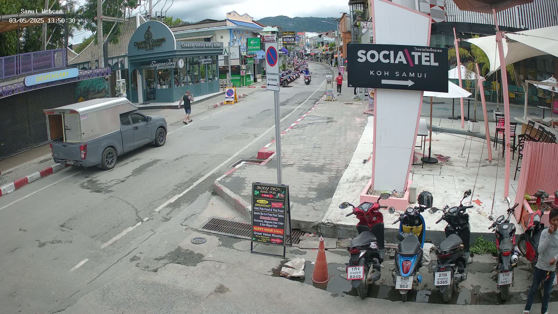 A street scene shows a gray pickup truck parked on the side of a road lined with shops and restaurants under an overcast sky, with several parked motorbikes and a few pedestrians.