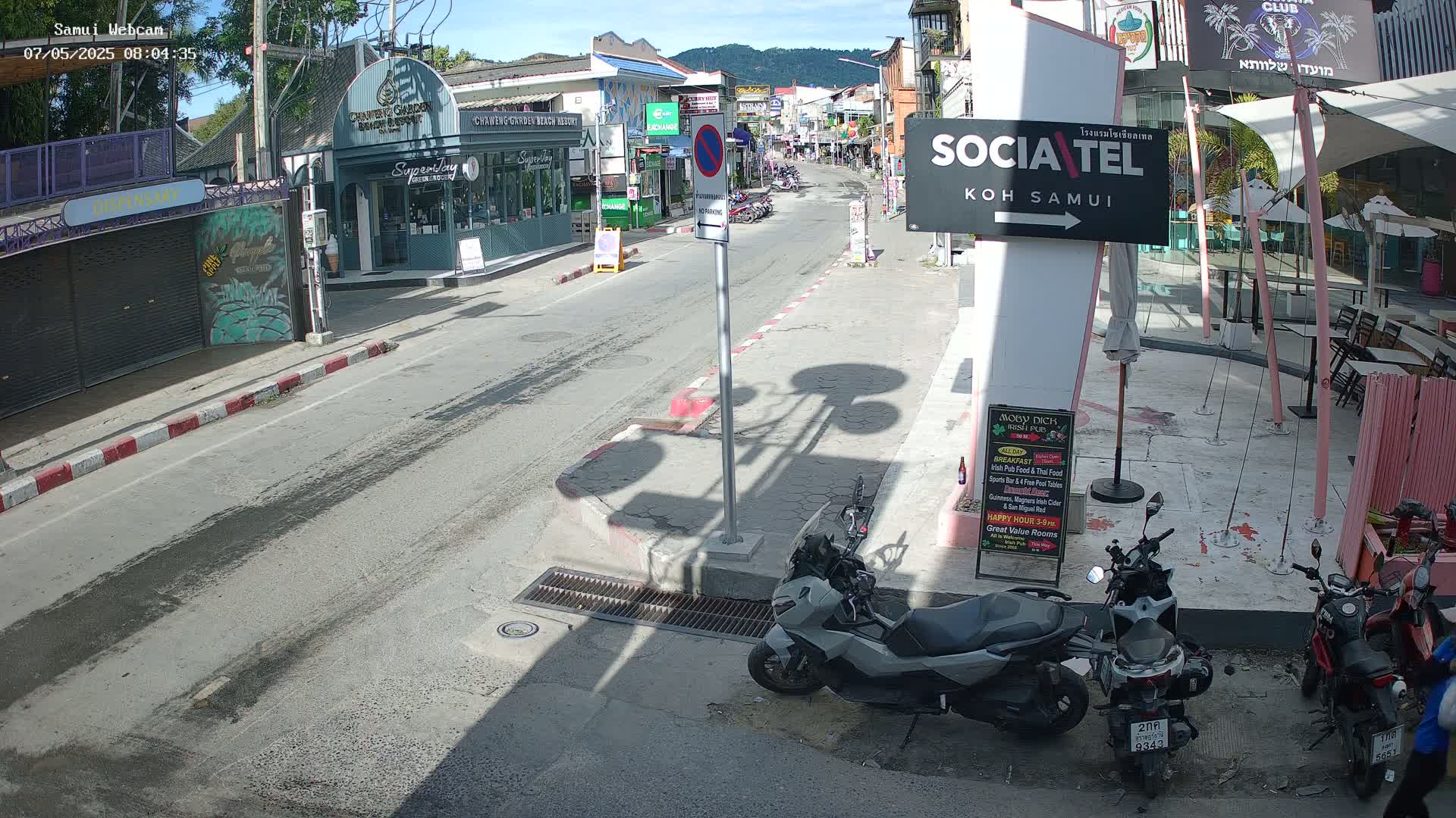 A sunny street scene shows parked motorbikes alongside buildings and a roadway with light traffic under a clear sky.