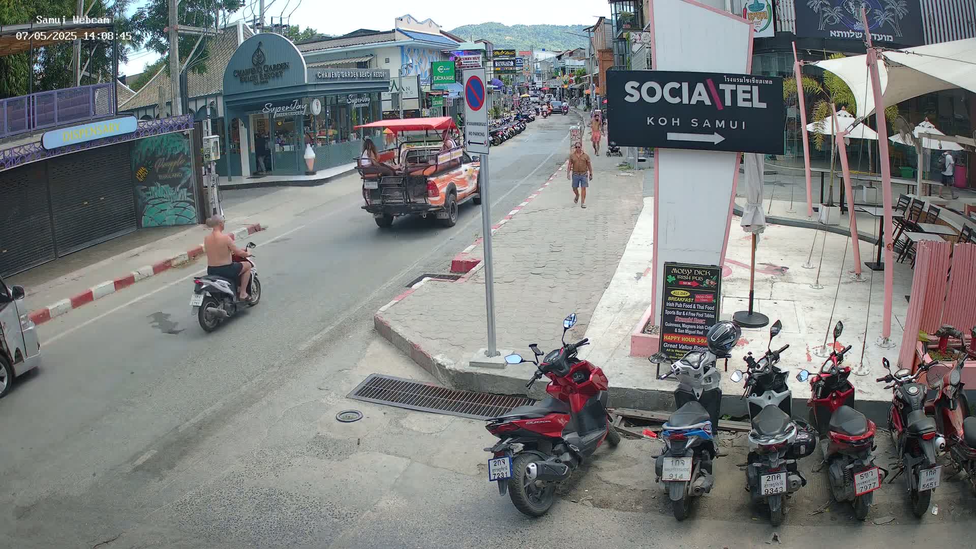 A sunny street scene shows a man on a scooter, two pedestrians, several parked motorbikes and scooters, a tourist vehicle, and shops and restaurants along the street.