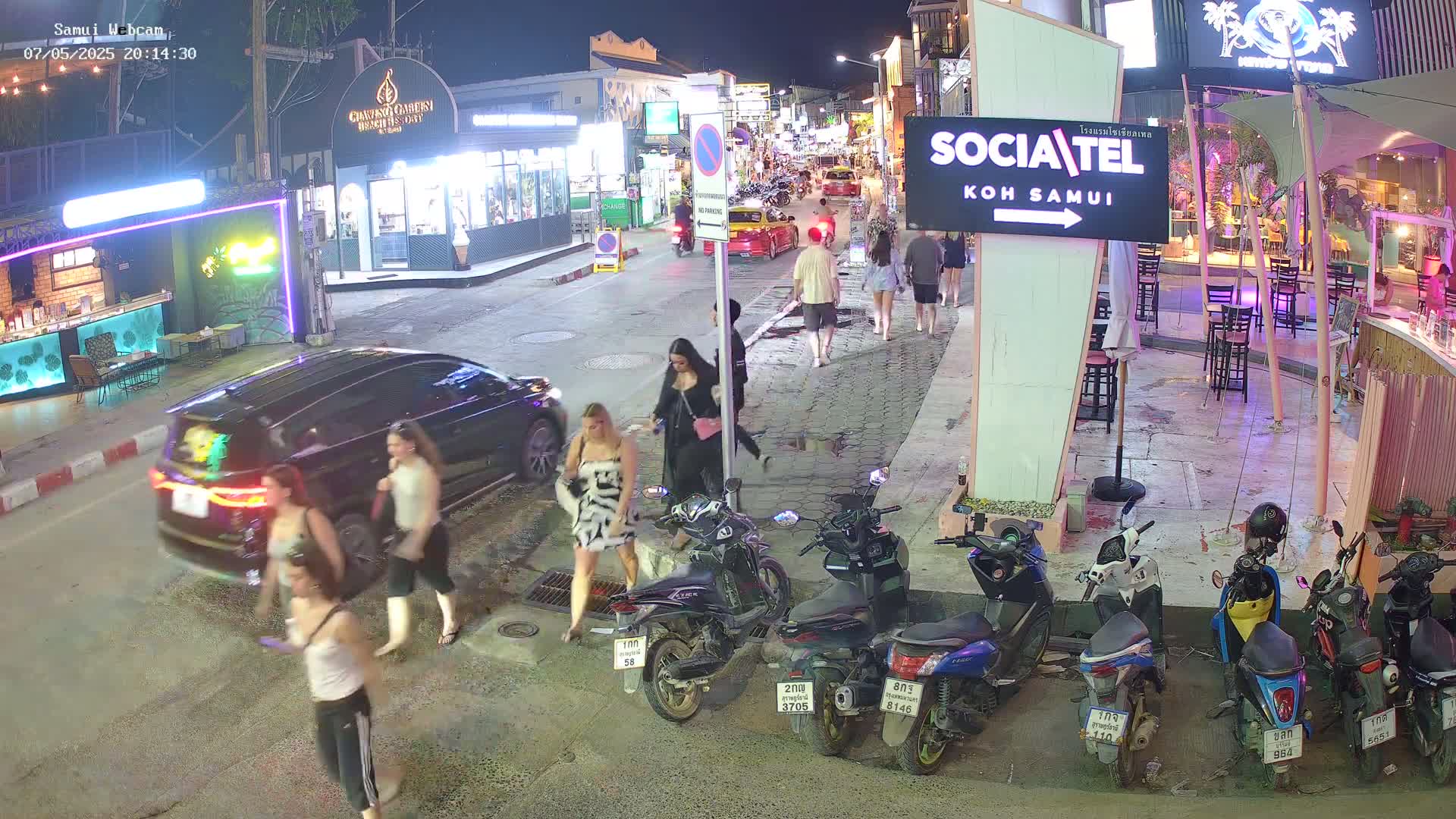 At night, several people walk along a street lined with shops and restaurants, near parked cars and motorbikes.
