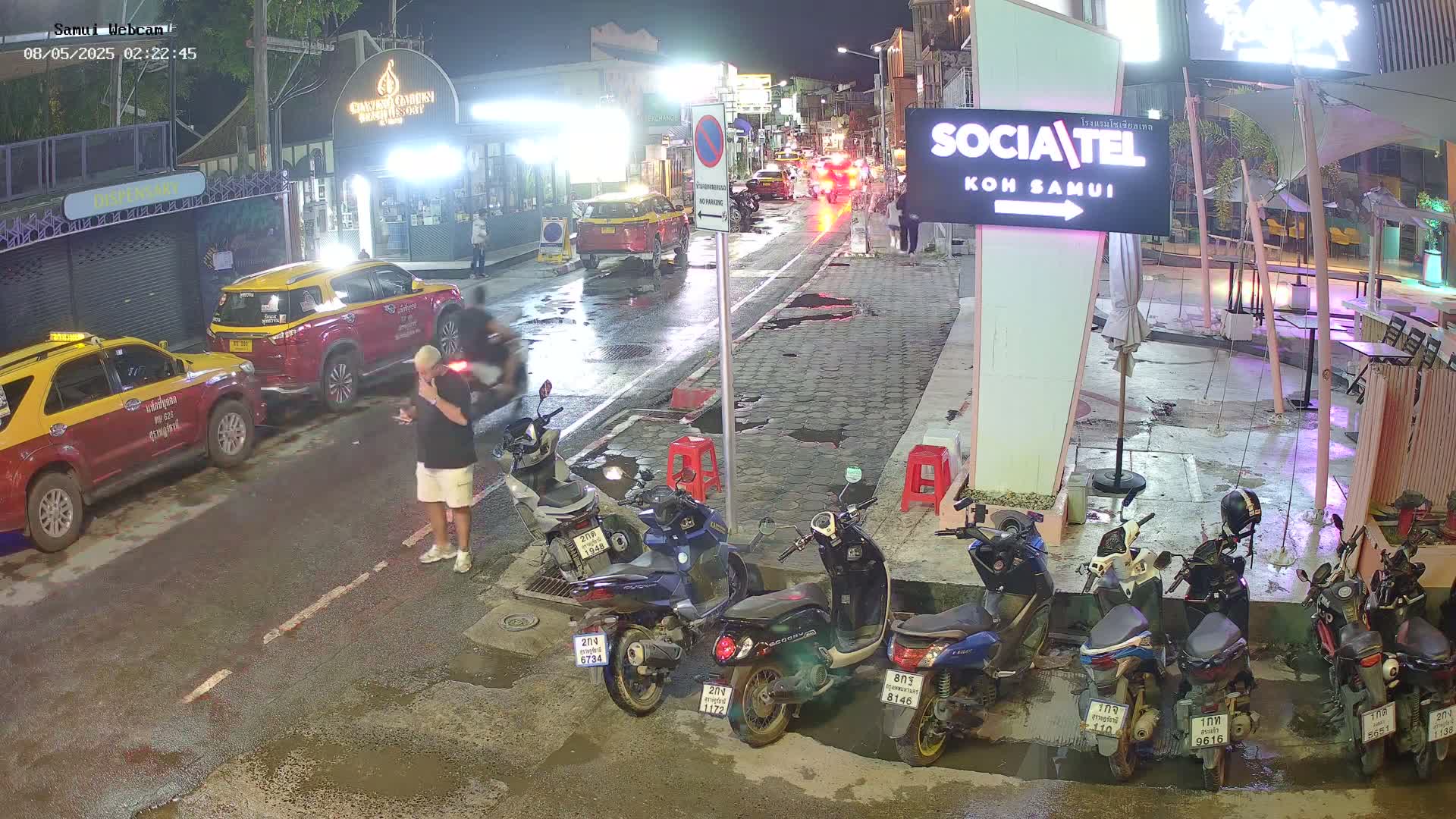 A nighttime street scene shows several parked motorbikes, two taxis, and a man standing near the curb under wet conditions.