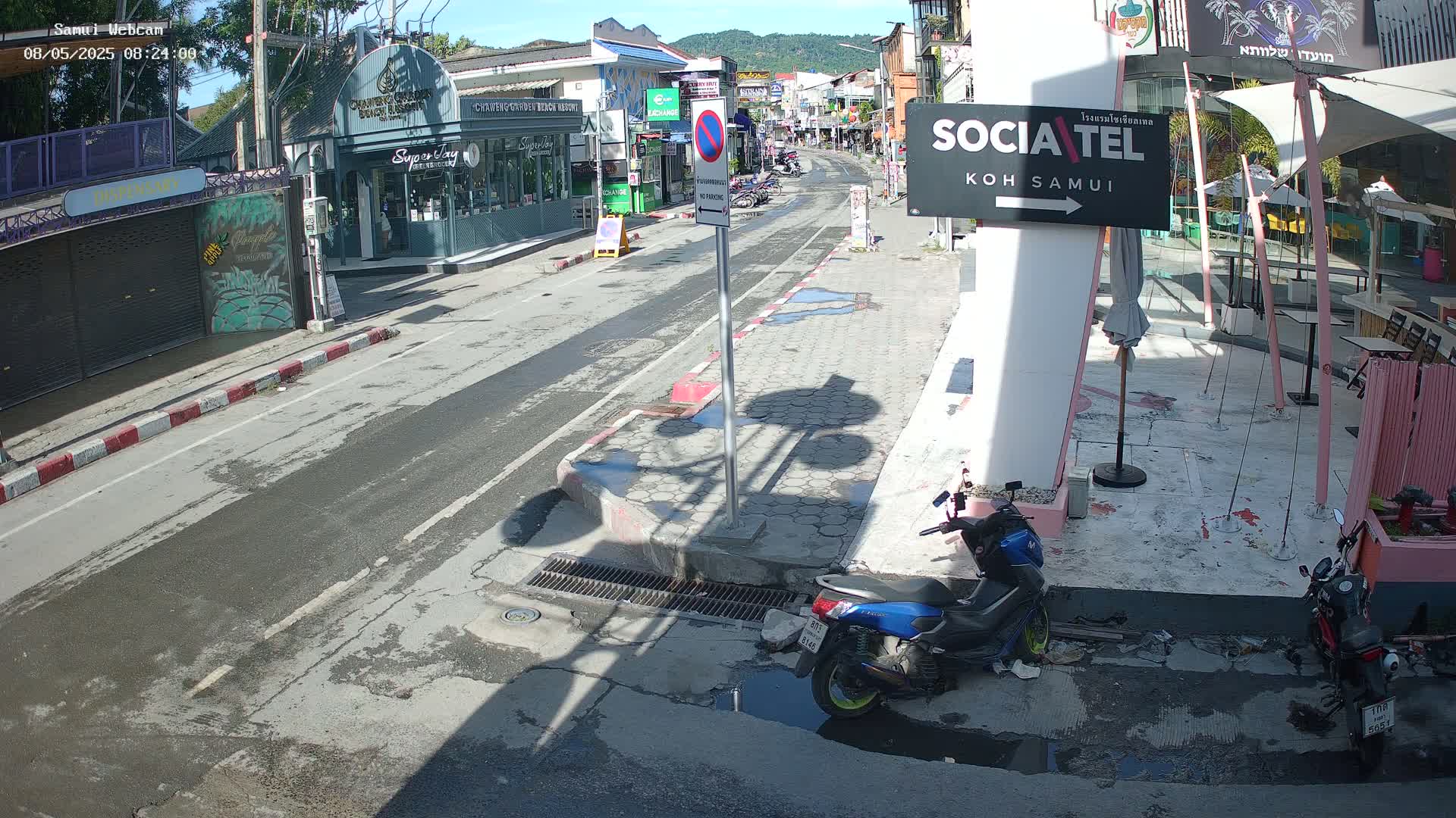 A sunny street scene shows a mostly empty road with parked motorcycles, shadows cast by buildings and a street light, and puddles of water on the pavement.