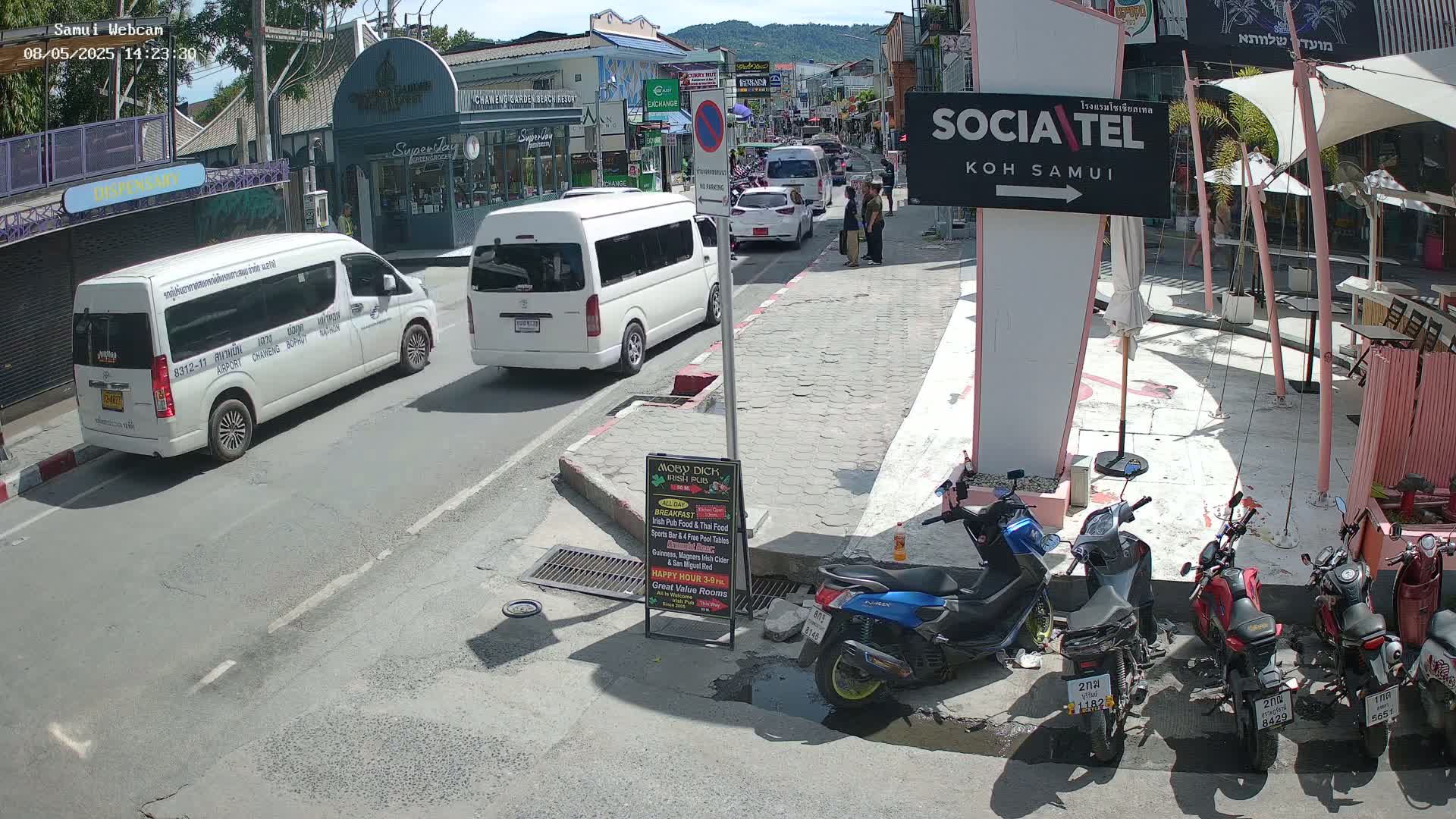 A sunny street scene shows several vehicles, including vans and motorbikes, parked alongside a roadway and in front of buildings.