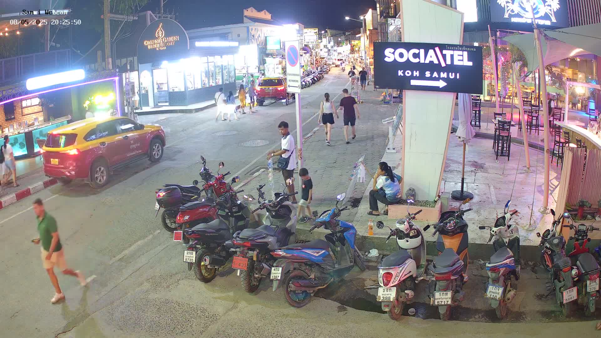 At night, a street scene shows pedestrians, parked cars and numerous motorbikes along a street lined with shops and restaurants under clear skies.