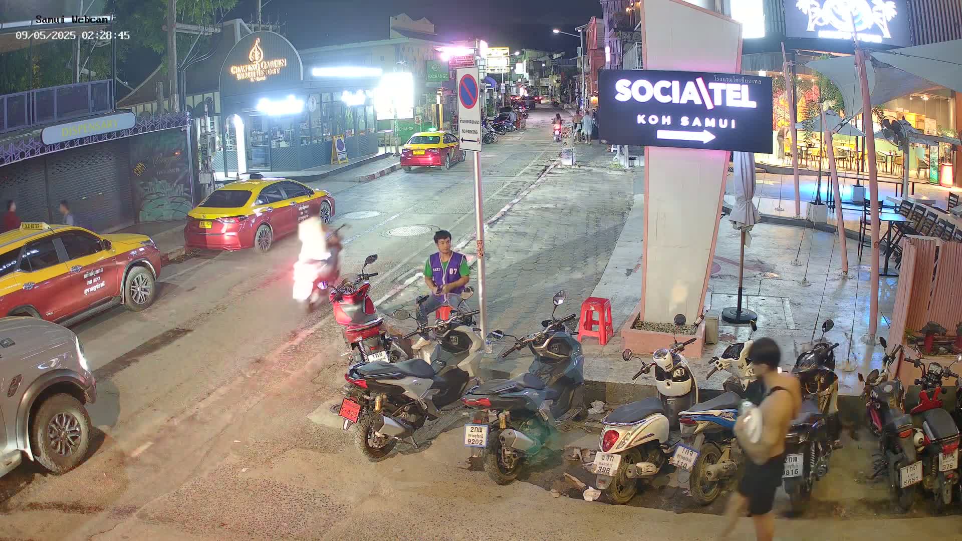 At night, a street scene shows several parked motorbikes and two taxis, with a man in a green vest sitting on a curb near a person walking away from the camera.