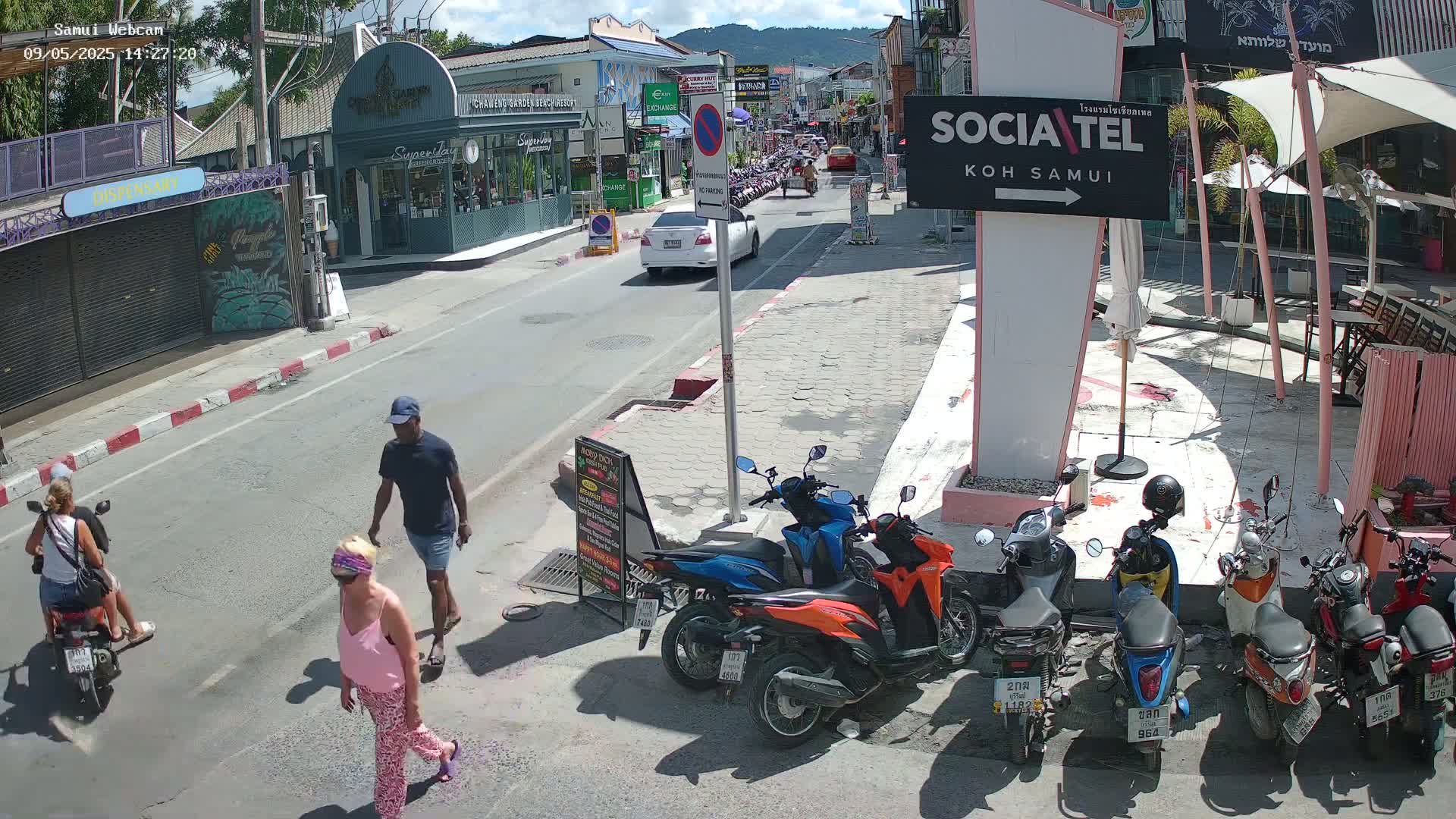 A sunny street scene shows several pedestrians and parked motorbikes along a roadway, with cars driving past shops and buildings.