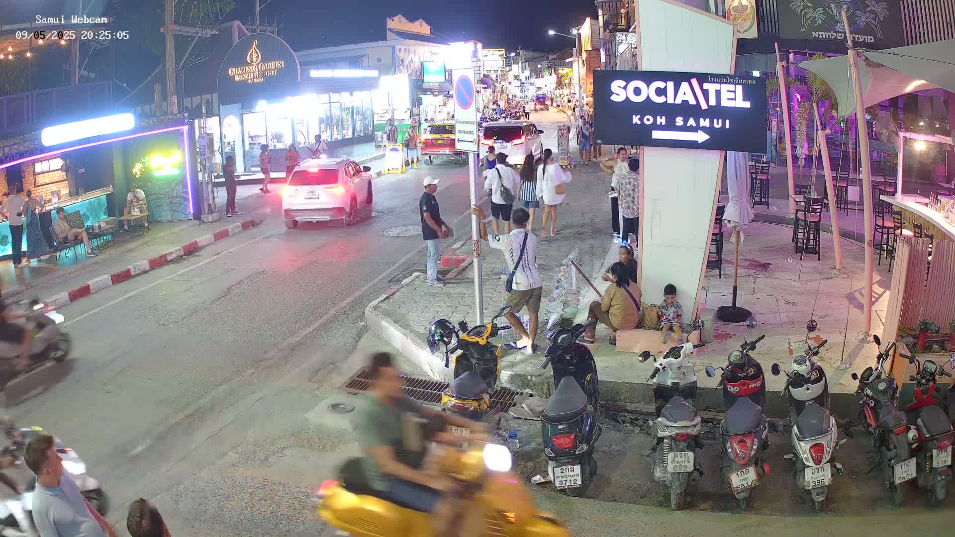 At night, a street scene shows people walking and sitting around parked motorbikes under ambient lighting.