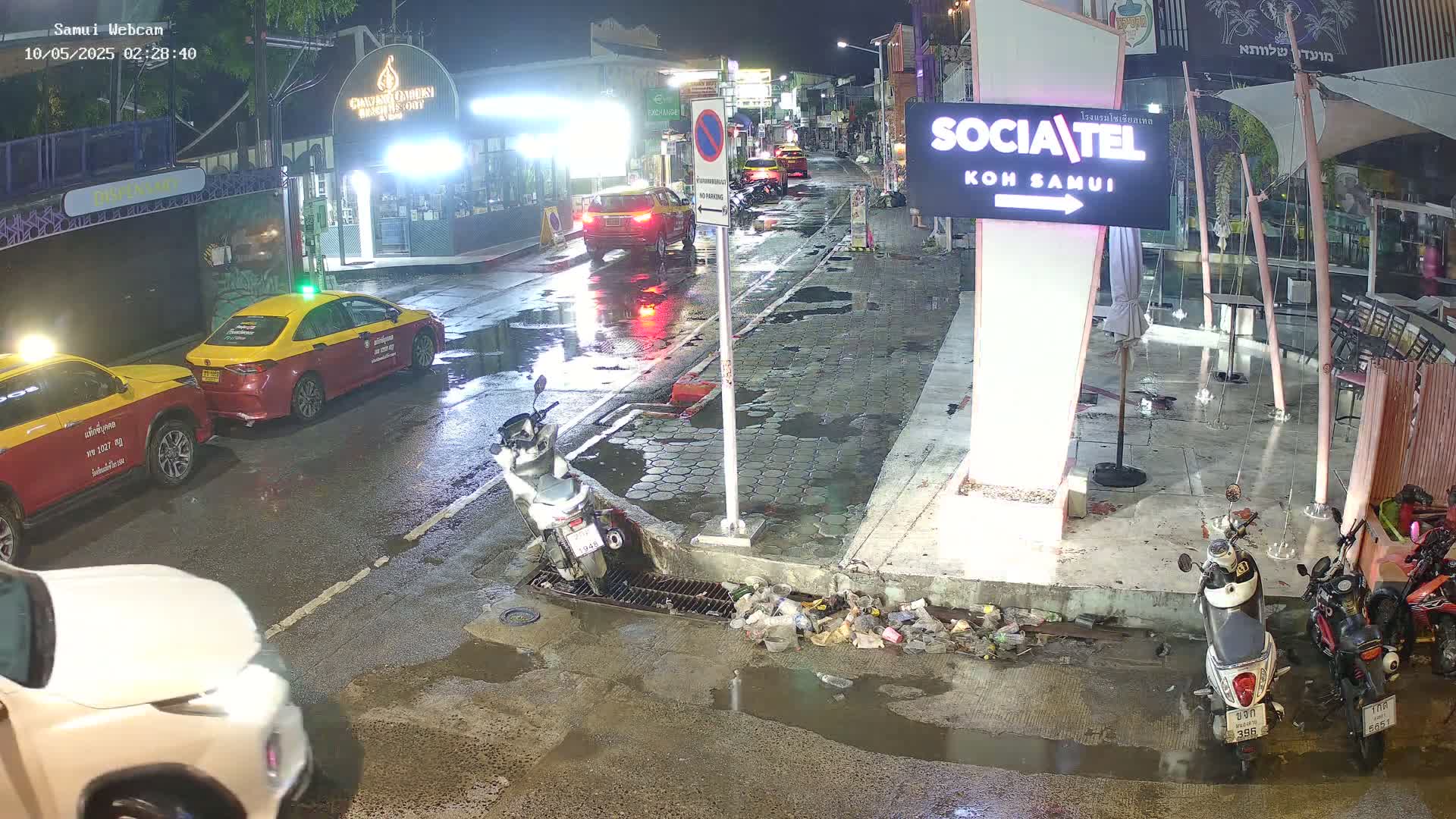 A wet, nighttime street scene shows several cars and motorcycles parked along the curb, with puddles of water reflecting the lights.