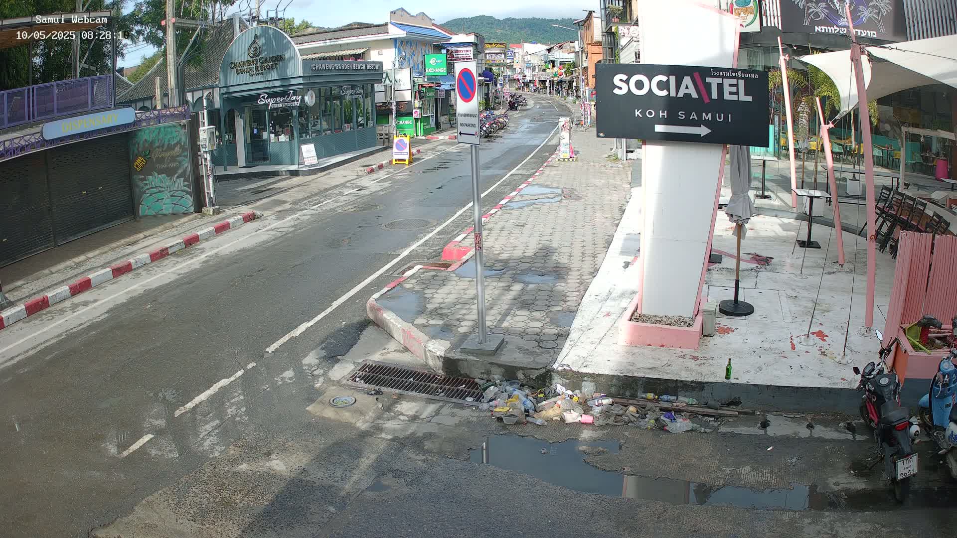 A mostly empty street scene on a partly sunny day shows puddles and some litter near the curb next to shops and restaurants.