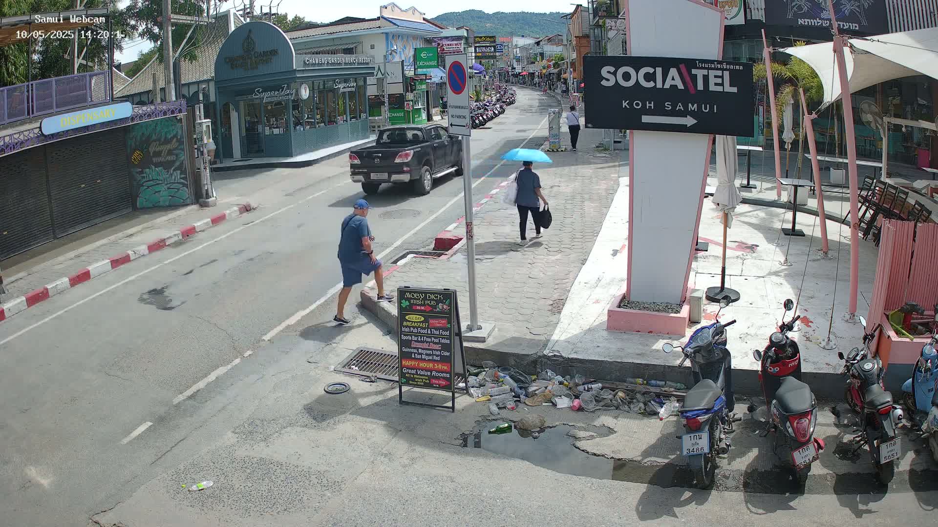 A sunny street scene shows pedestrians walking, a pickup truck parked, and several mopeds parked, with some litter on the ground near a restaurant sign.