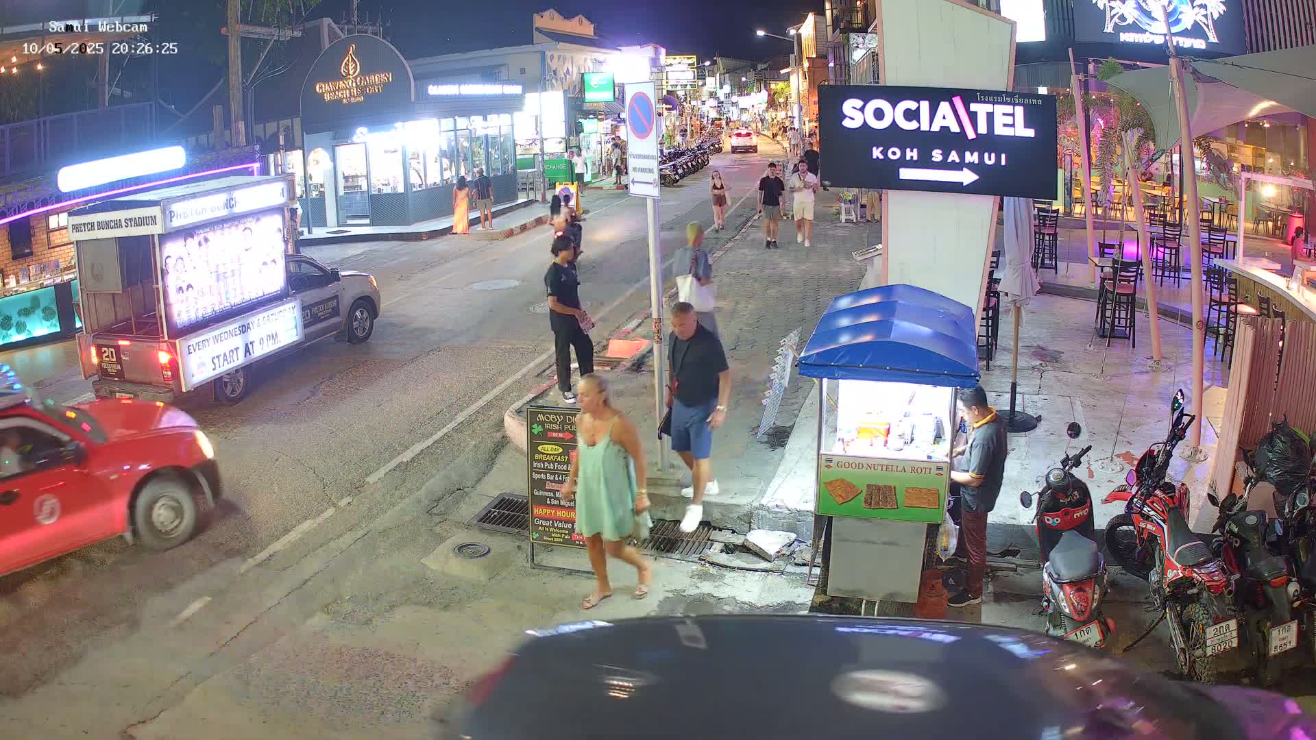 A nighttime street scene shows several people walking past shops and food vendors, with parked cars and motorcycles nearby.