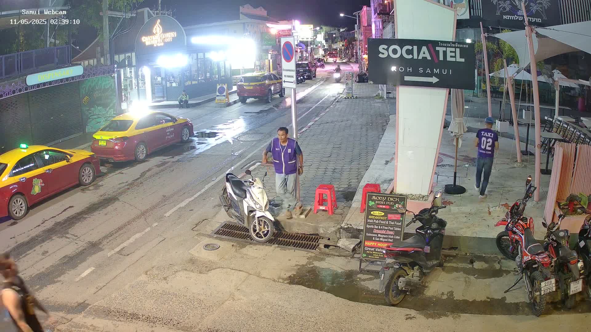 A night scene on a city street shows several parked cars, a man standing near a scooter, and several motorcycles parked near a restaurant under a partially cloudy sky.