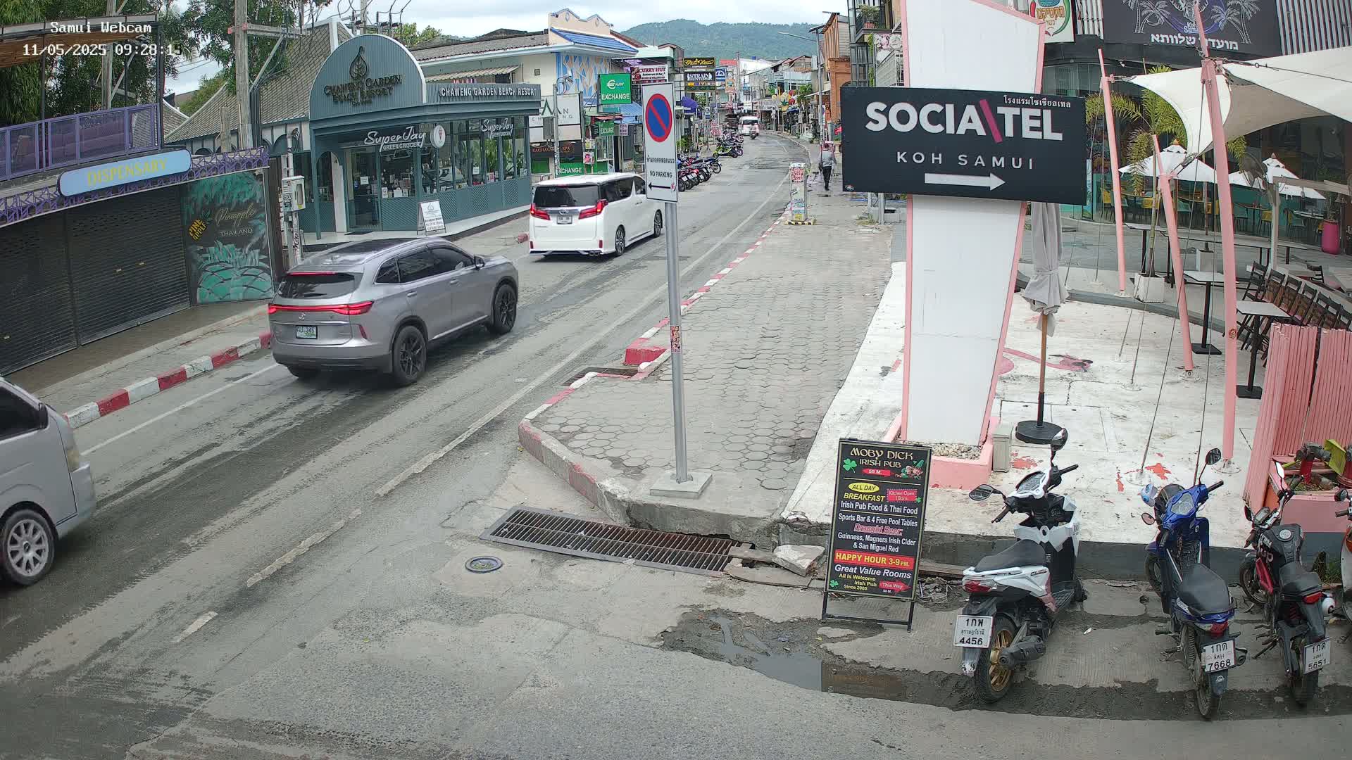 A street scene shows several cars and scooters parked along a road, with buildings and outdoor seating areas visible on either side, under a mostly sunny sky.